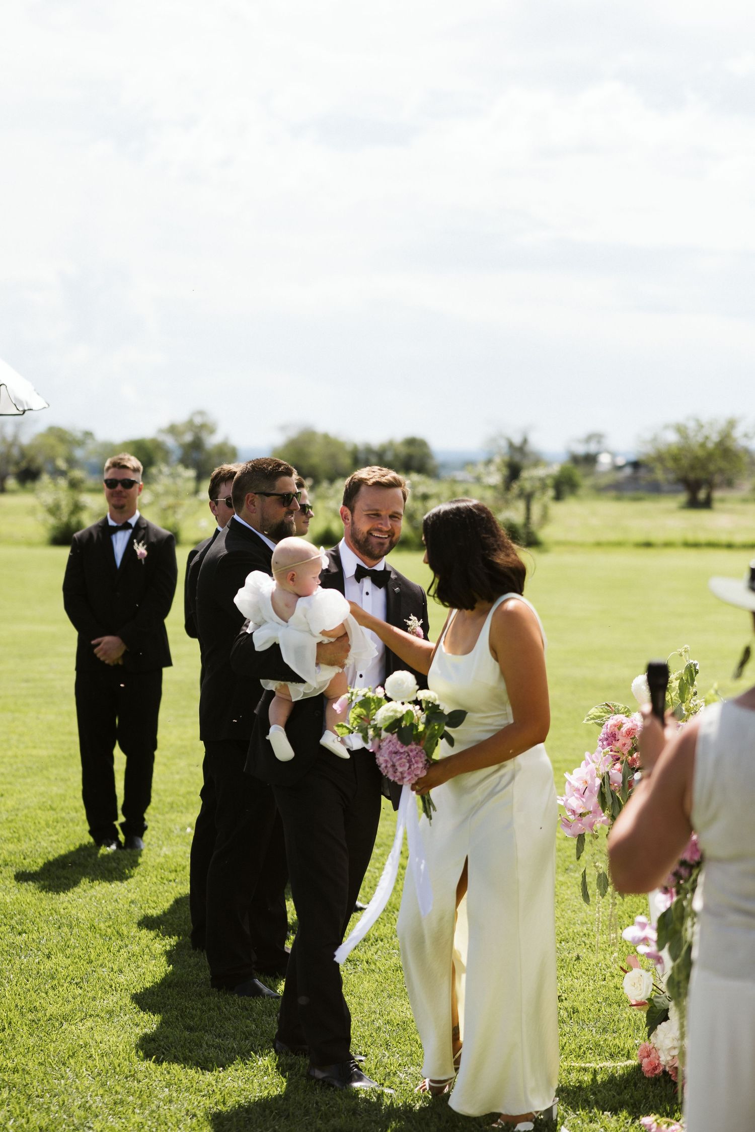 Wedding ceremony takes place on a sunny lawn with attendants and guests gathered around.