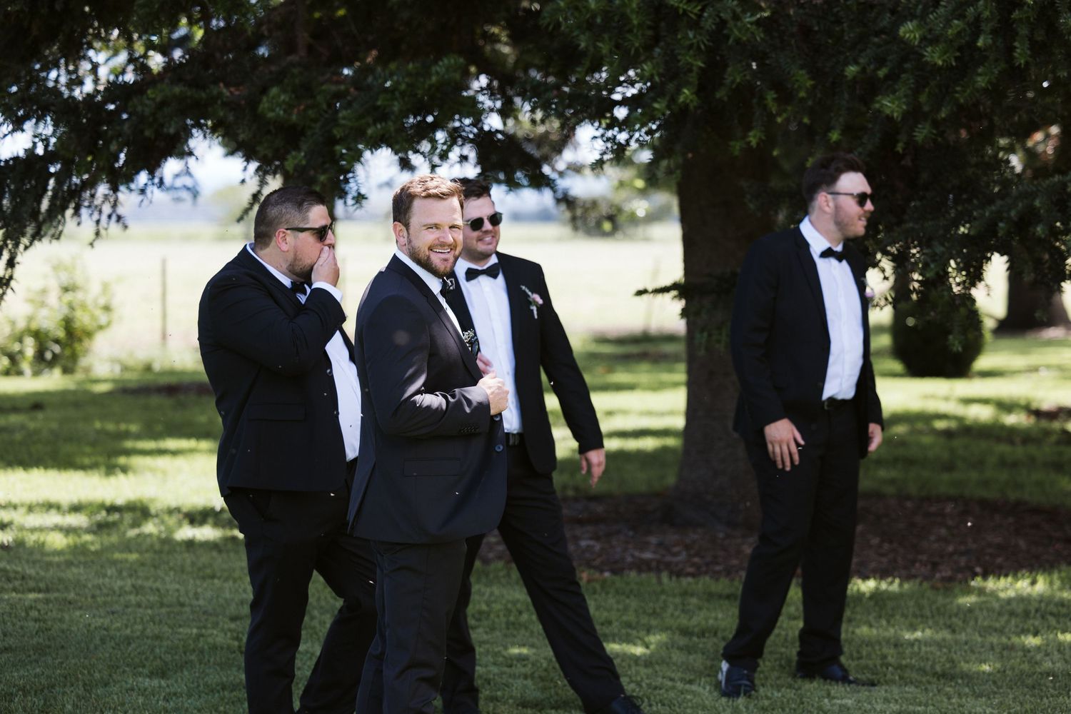 Groomsmen in formal attire walking and laughing on garden lawn.