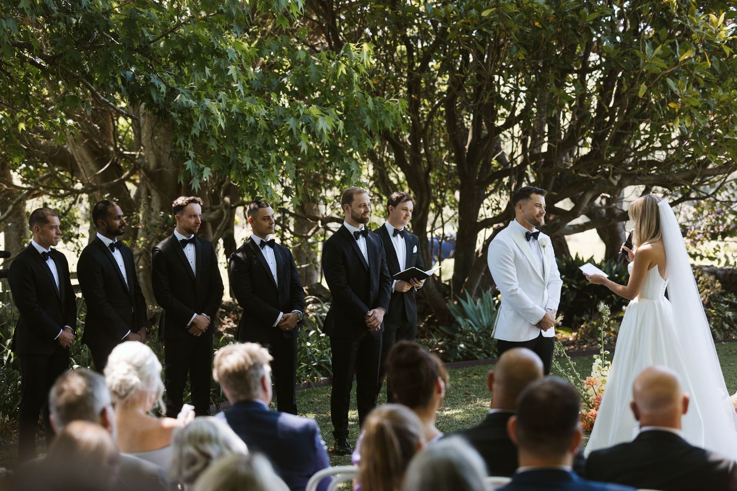 Wedding ceremony in progress with wedding party and guests in garden setting.