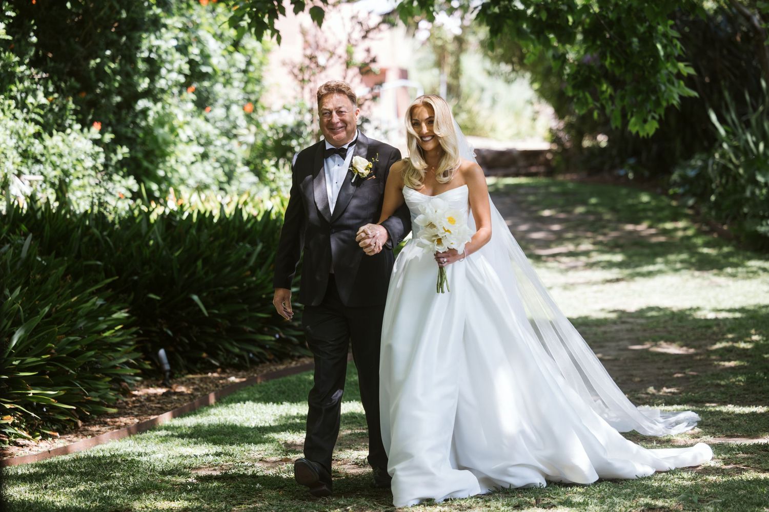 Couple poses in formal wedding attire under trees in garden setting.
