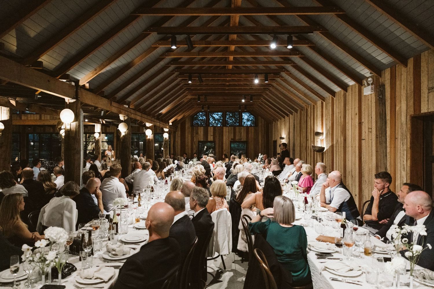 Wedding reception in a vaulted wooden hall with guests dining at elegantly set tables.