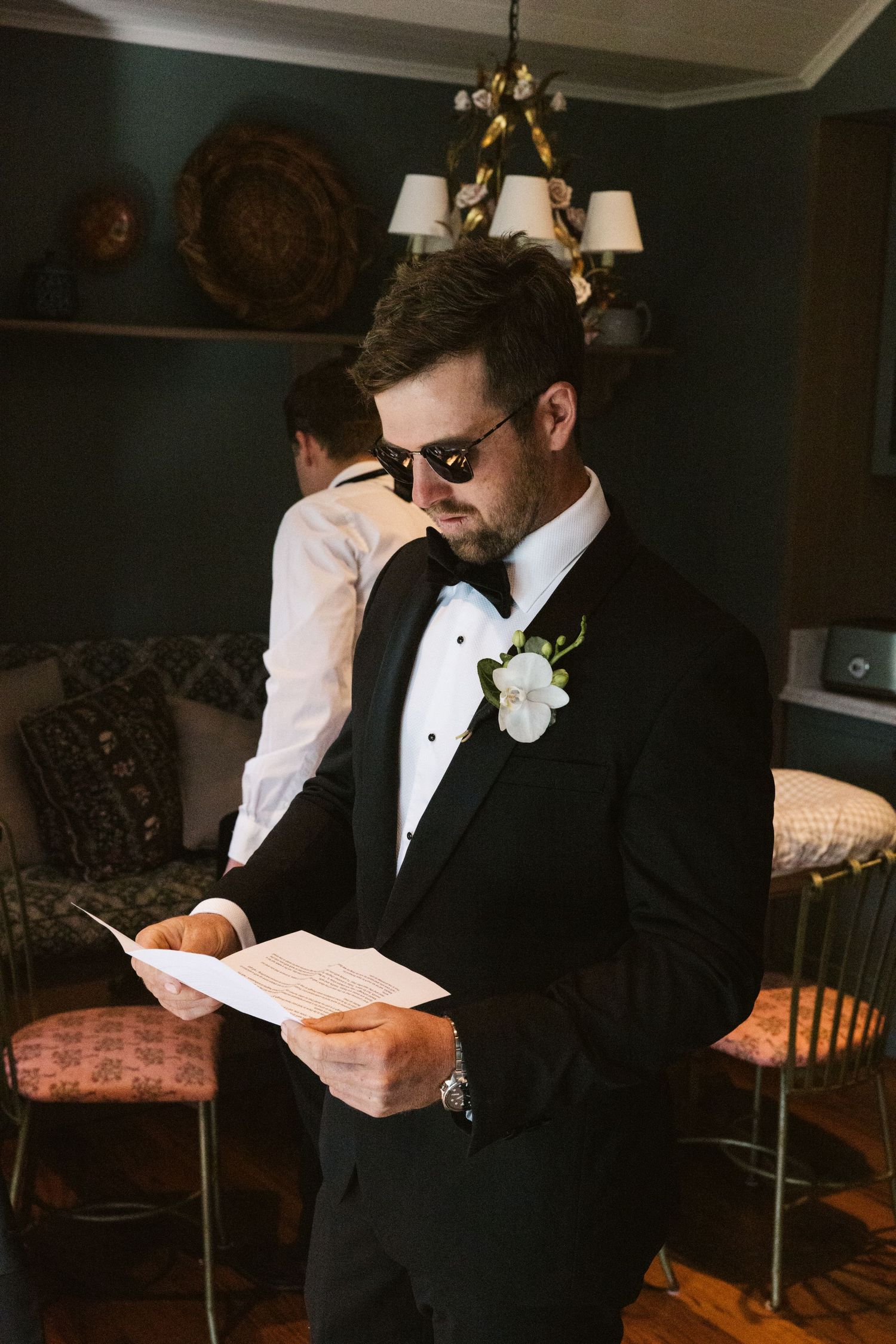 Groom in tuxedo and sunglasses reading document in elegantly furnished room.