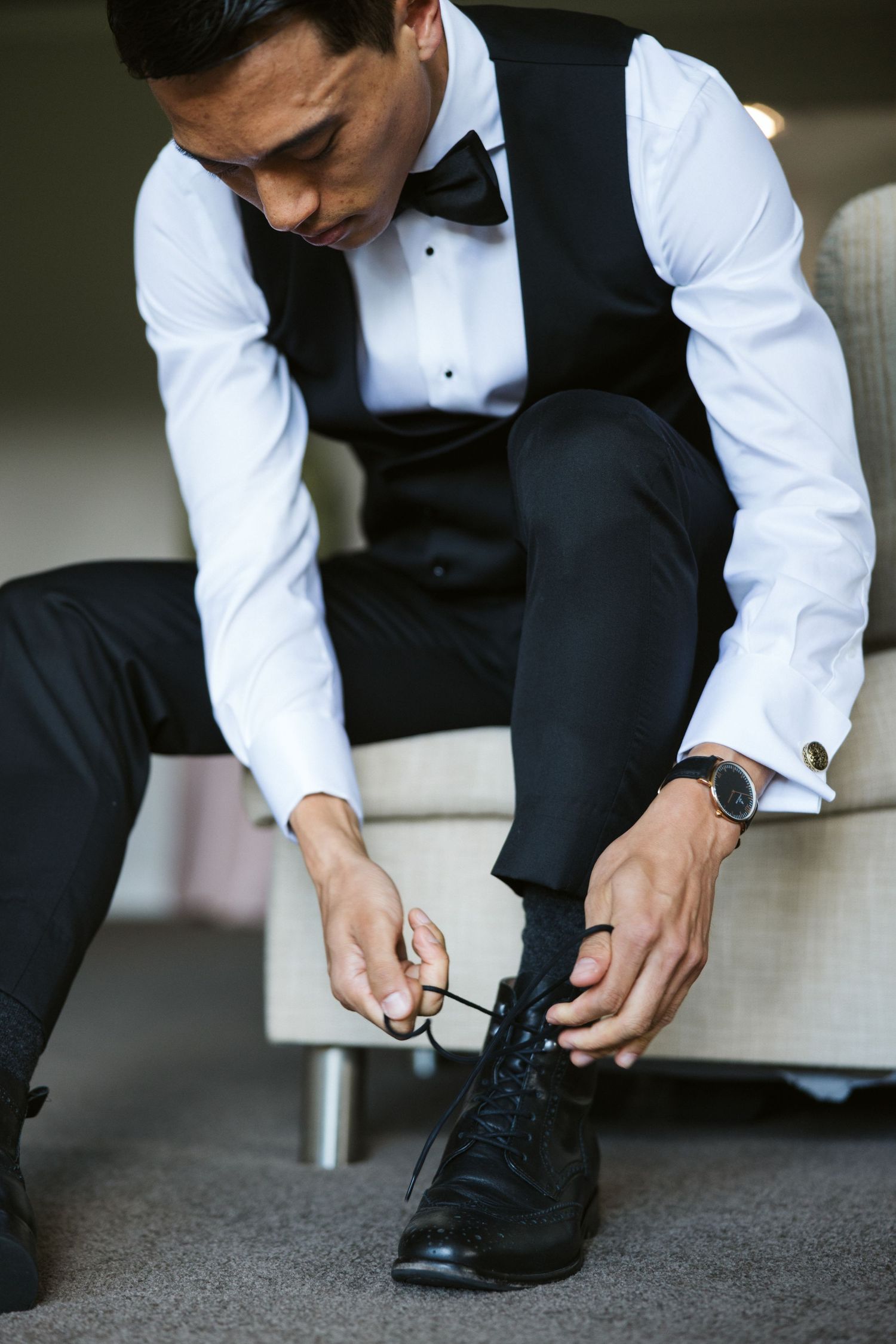 A formally dressed person ties their black dress shoes while wearing a tuxedo vest and dress shirt.