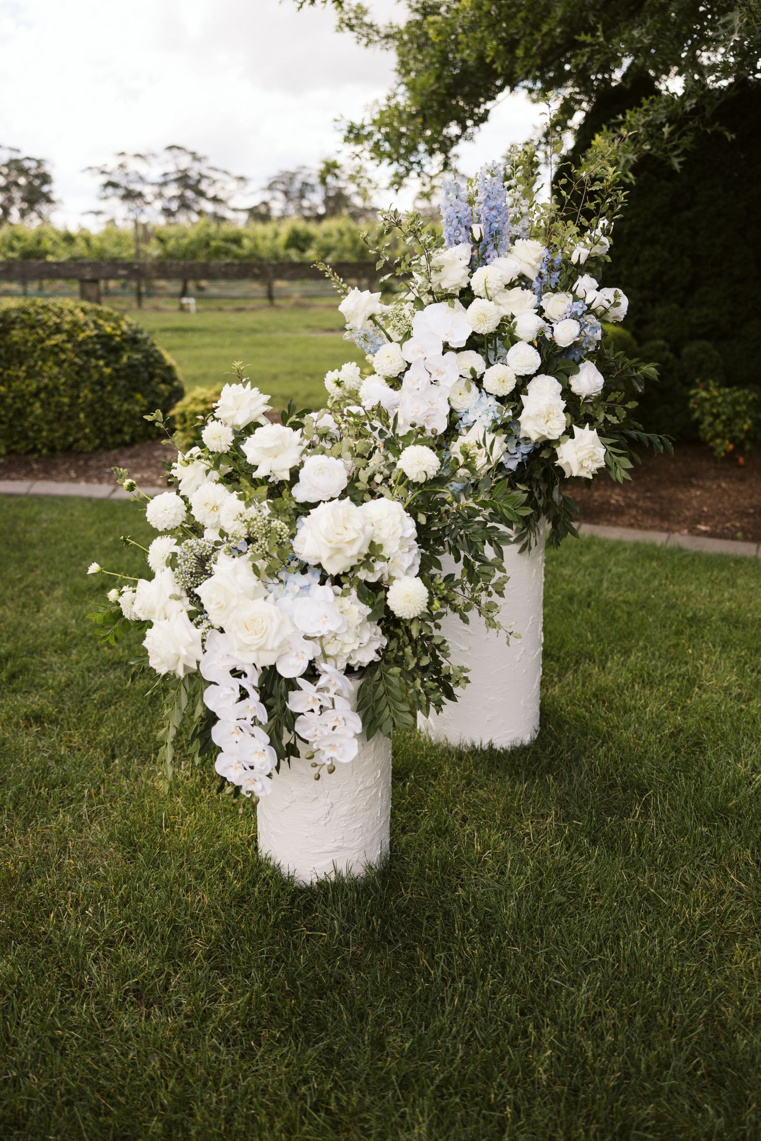 Elegant white floral arrangements in cylindrical white vases placed on a grassy lawn.