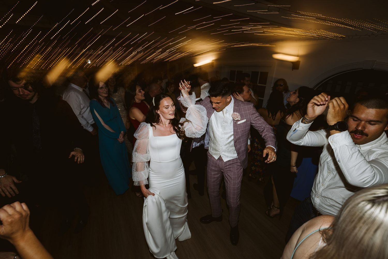 Bride and groom dance joyfully with guests at a lively wedding reception party under warm lights.