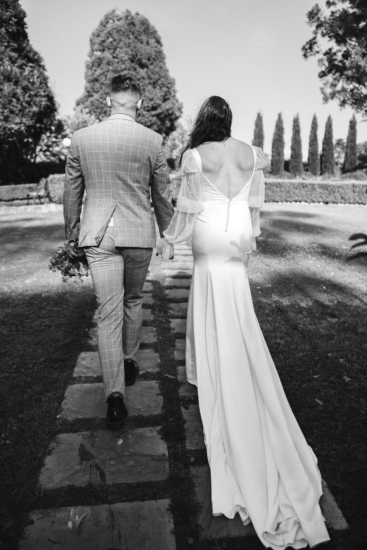Black and white photo of bride and groom walking hand in hand along stone path, bride in backless gown, groom in plaid suit.