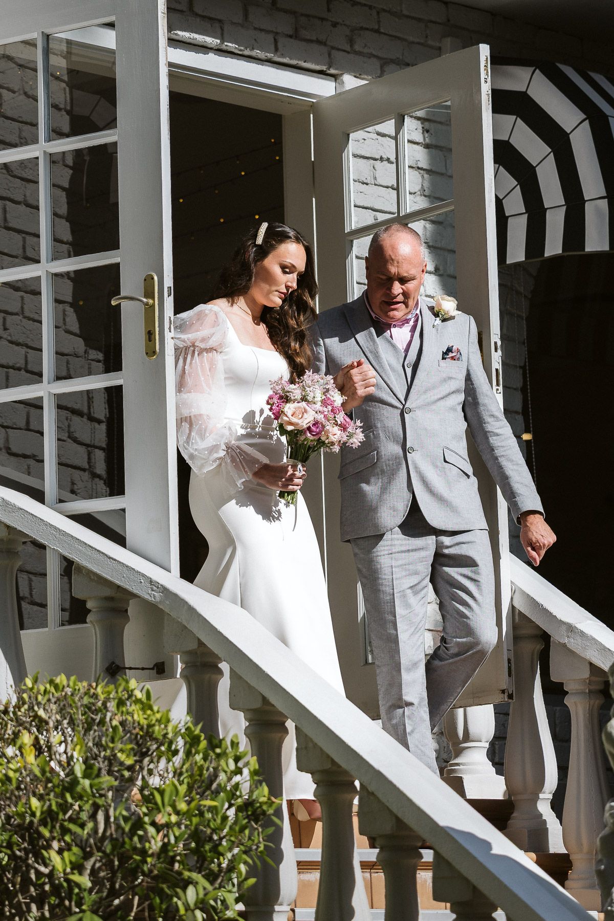 Bride in white gown holding bouquet walks down steps with older man in gray suit at wedding venue.