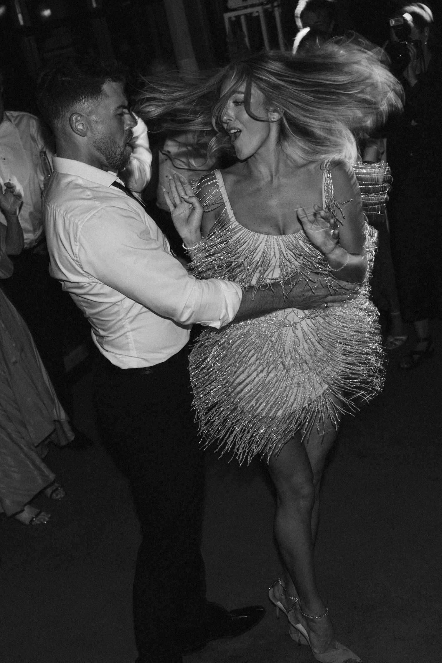A couple dances energetically on a dark dance floor with the woman wearing a shimmering fringe dress.