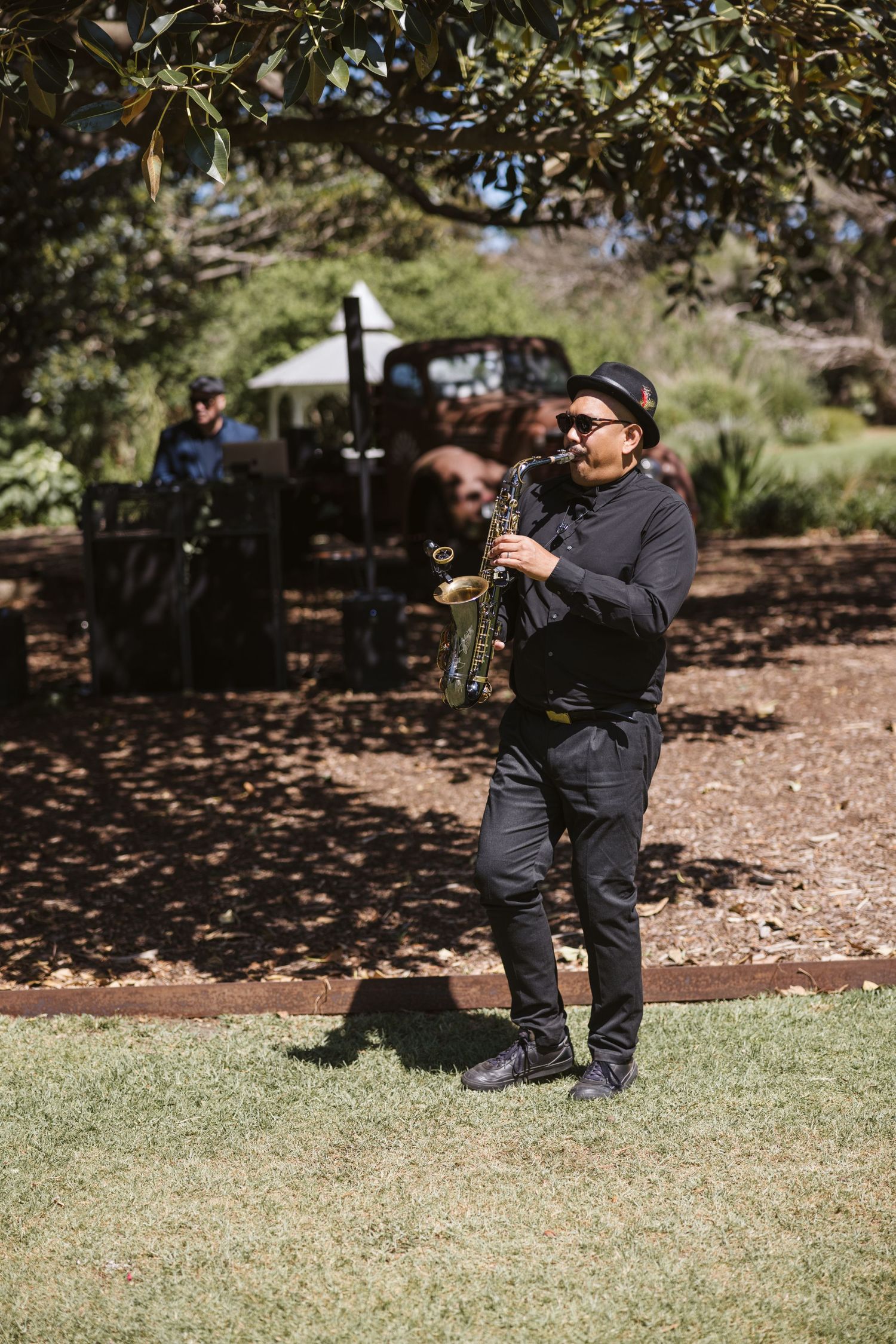 Saxophone player performing in casual attire under trees at outdoor event.
