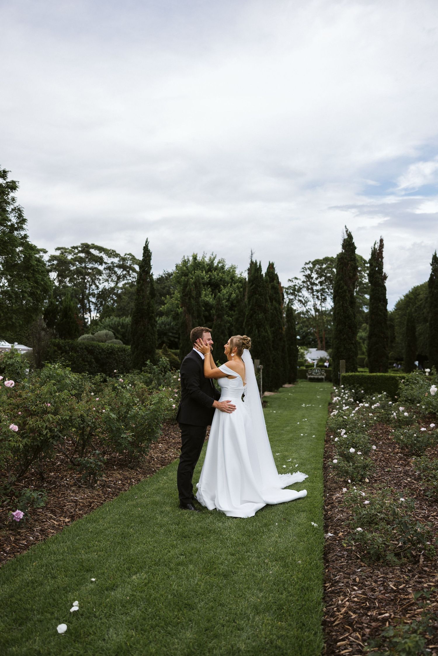 Wedding couple embracing on a manicured lawn path lined with cypress trees.