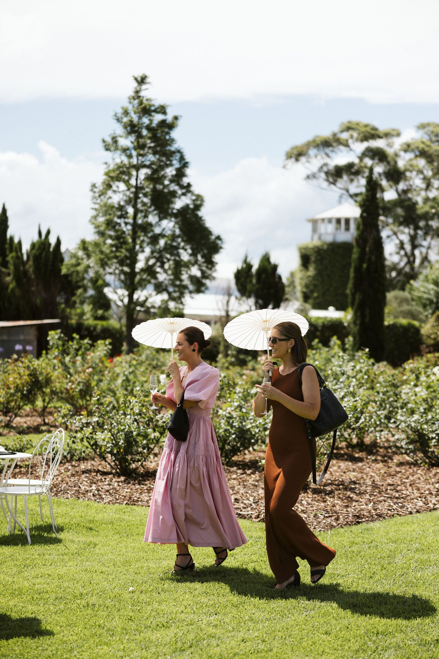 Two guests with parasols walking through garden during sunny outdoor event.