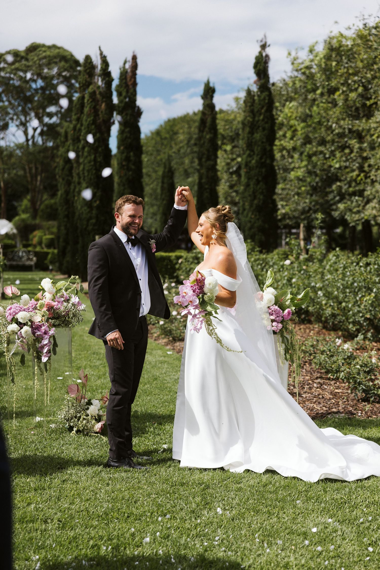 Joyful wedding moment in garden with cypress trees and pink floral arrangements.