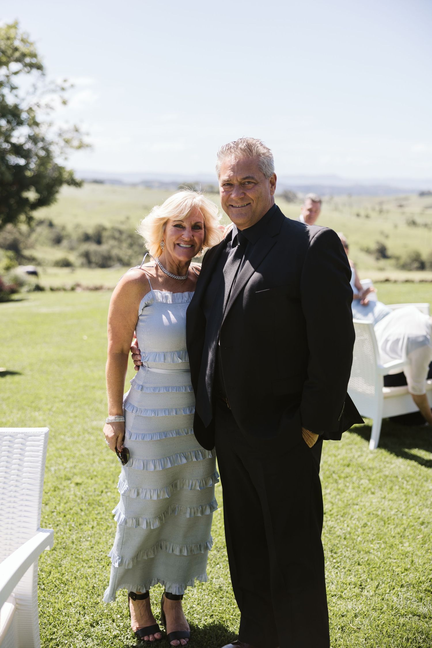 Formally dressed couple posing together on a sunny lawn with scenic views in the background.