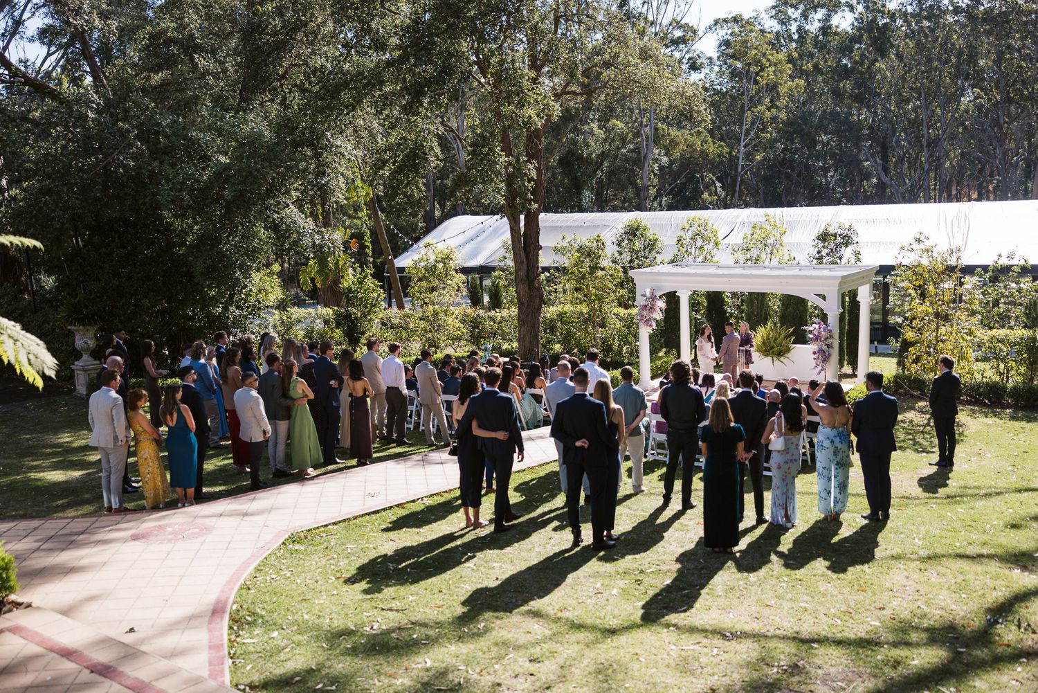 Guests gathered outdoors for a wedding ceremony in a lush garden setting with a white pergola.