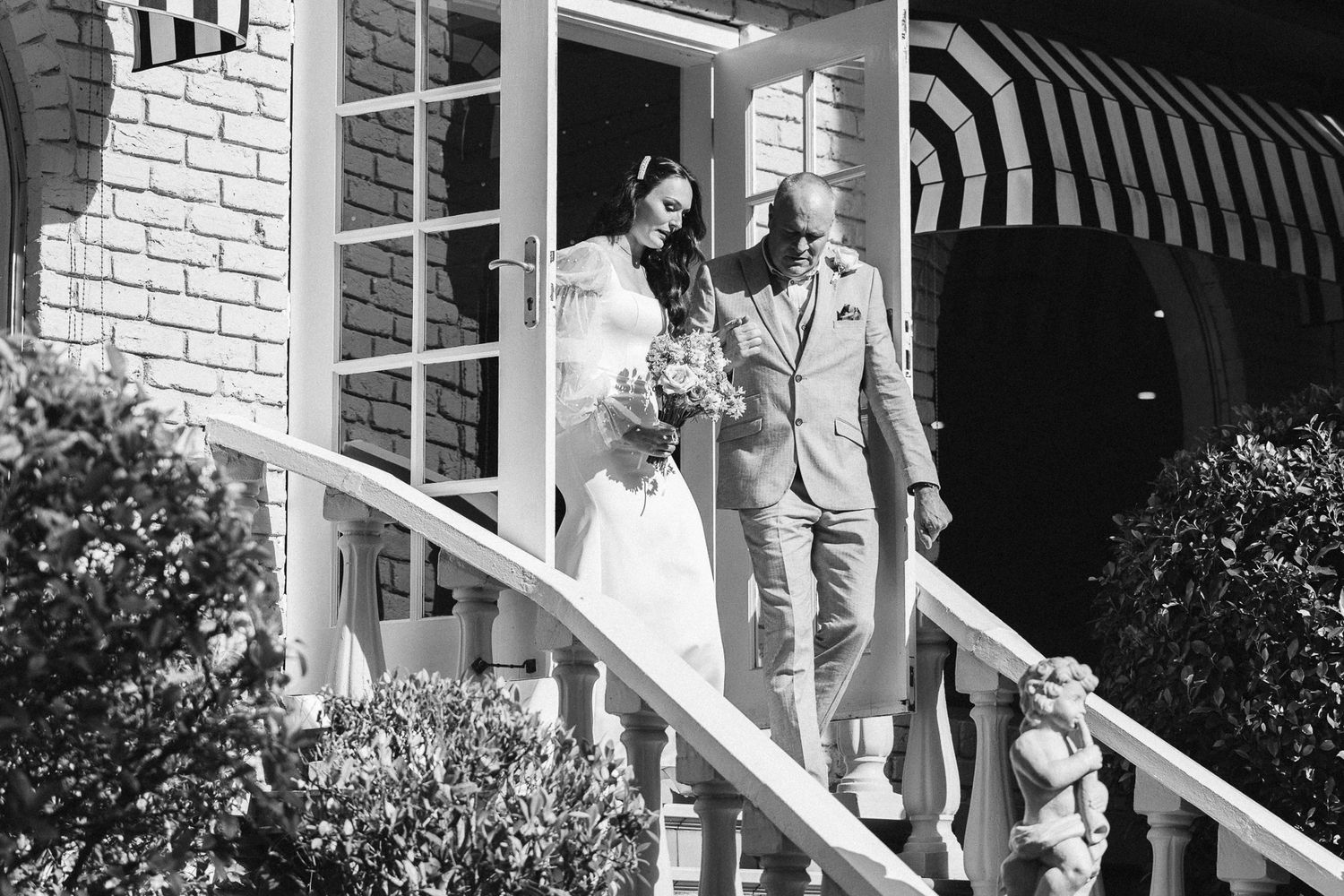 Black and white photo of a bride and groom descending elegant porch stairs at a classic venue on their wedding day.