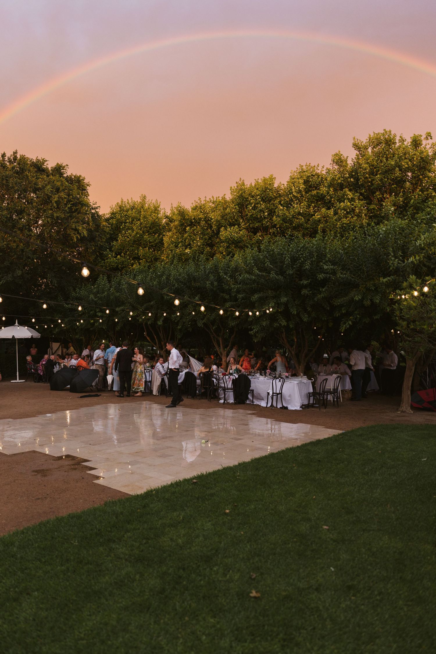 Outdoor wedding reception with string lights and rainbow in the sky above a dance floor.