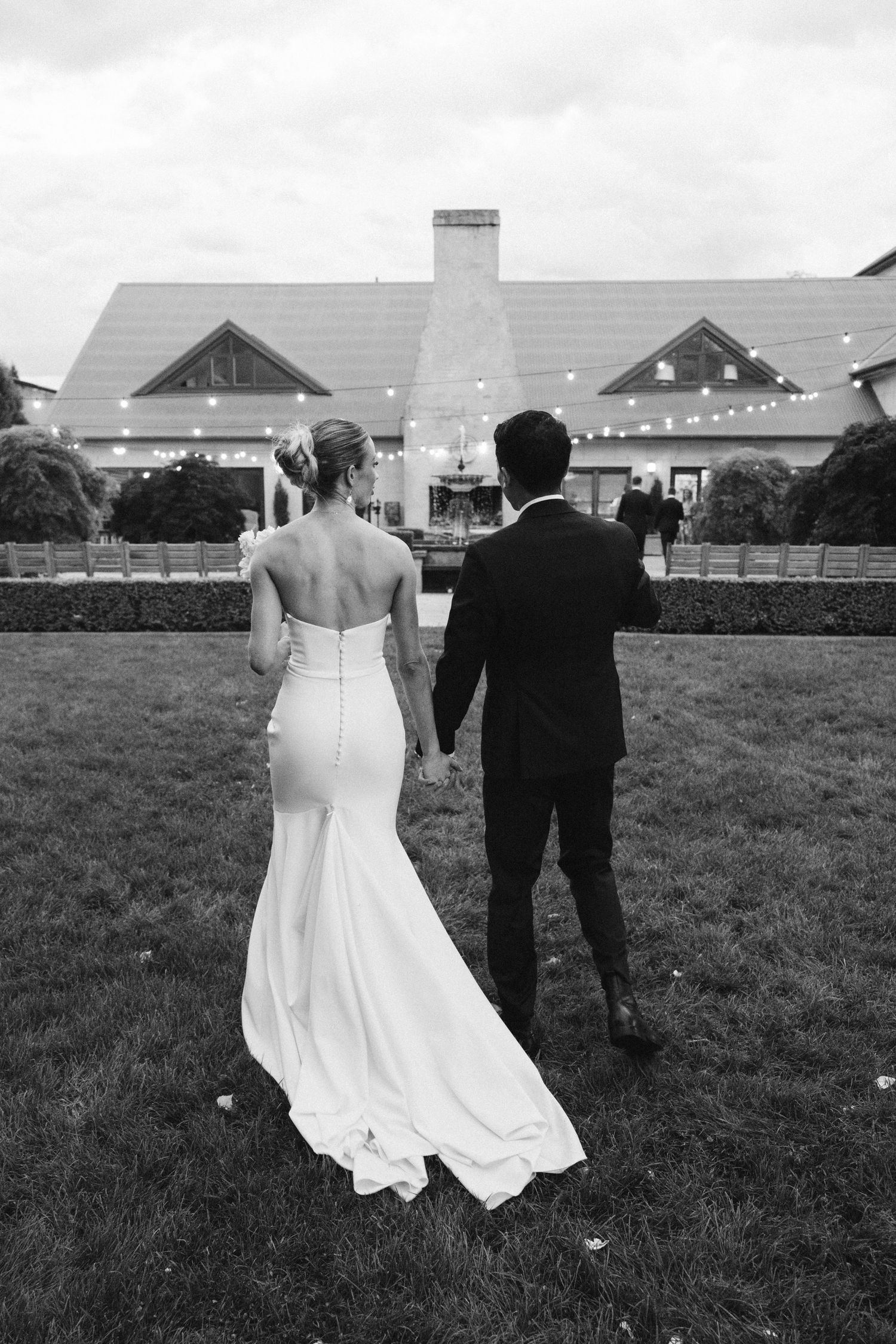 A couple in wedding attire standing on a lawn admiring a grand house with gabled roof in black and white.