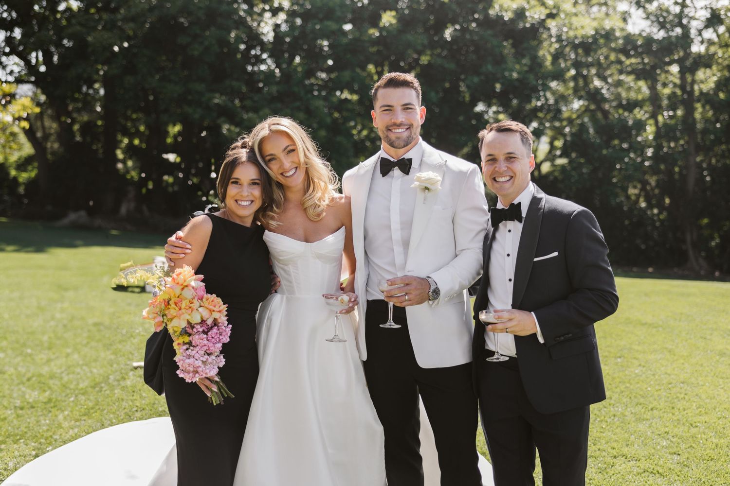 Wedding party poses together on grass with bride in white dress and attendants in dark attire.