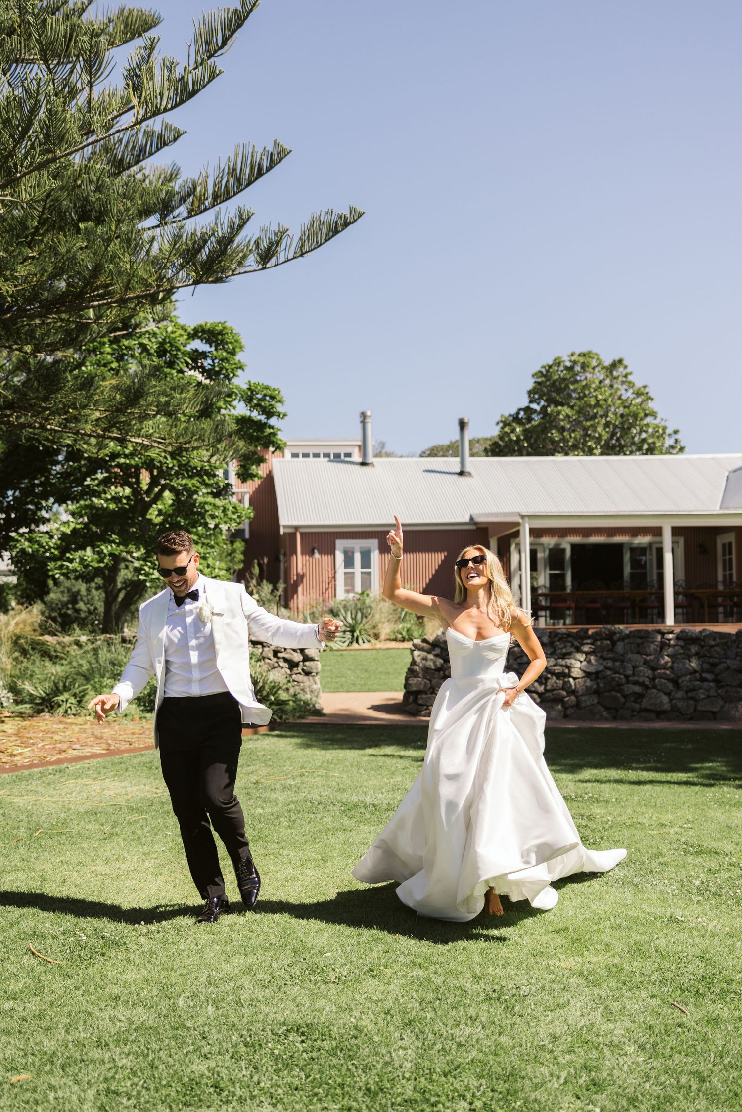 Couple dancing playfully on the lawn in front of a brown house with stone wall.