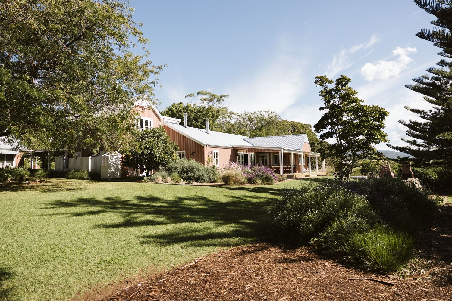Pink and white country house with wraparound porch nestled among manicured gardens and pine trees.