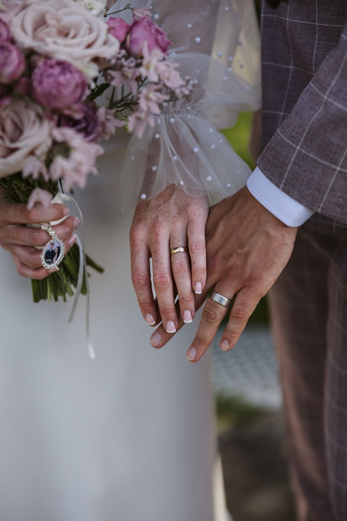 Newlyweds displaying wedding bands on joined hands, with bride holding pink floral bouquet and sapphire pendant.