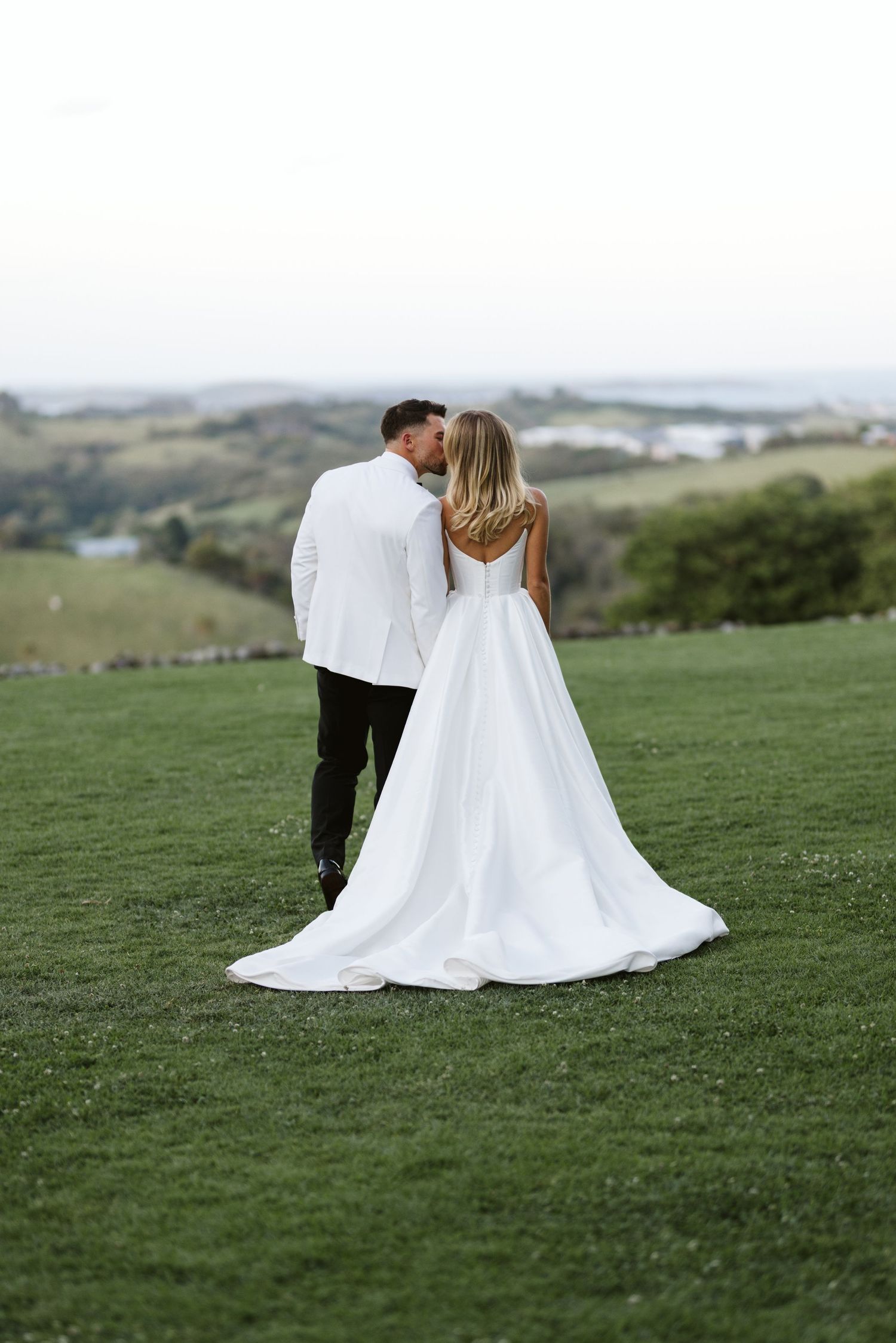 A couple in wedding attire embrace while standing on a hilltop overlooking a scenic landscape.