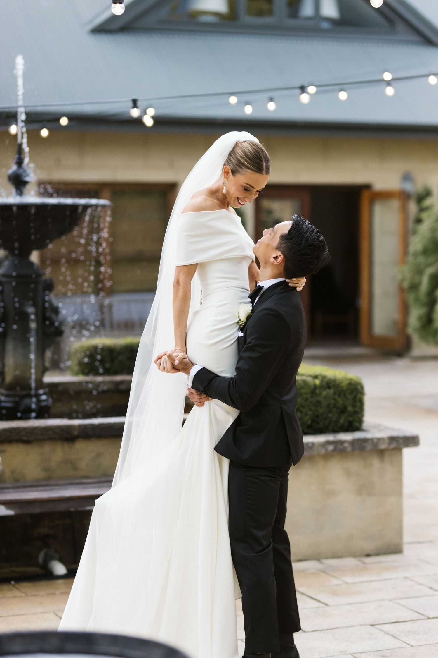 Intimate moment between newlyweds near fountain with string lights in background.