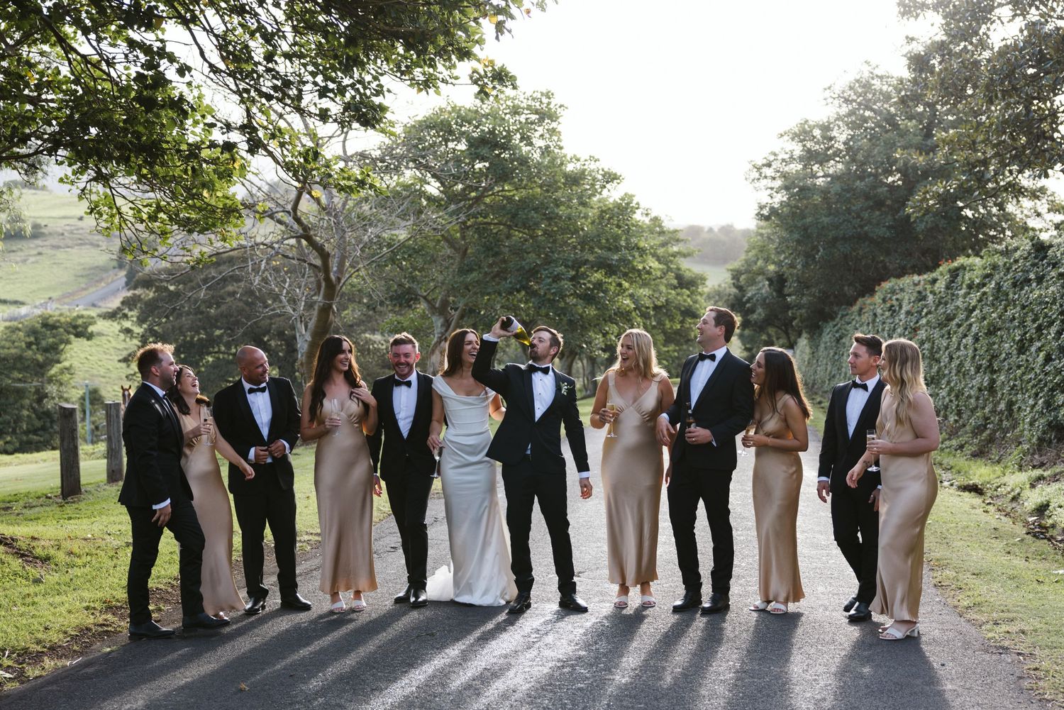 Wedding party posing together on a scenic rural road with trees and hedges in the background.