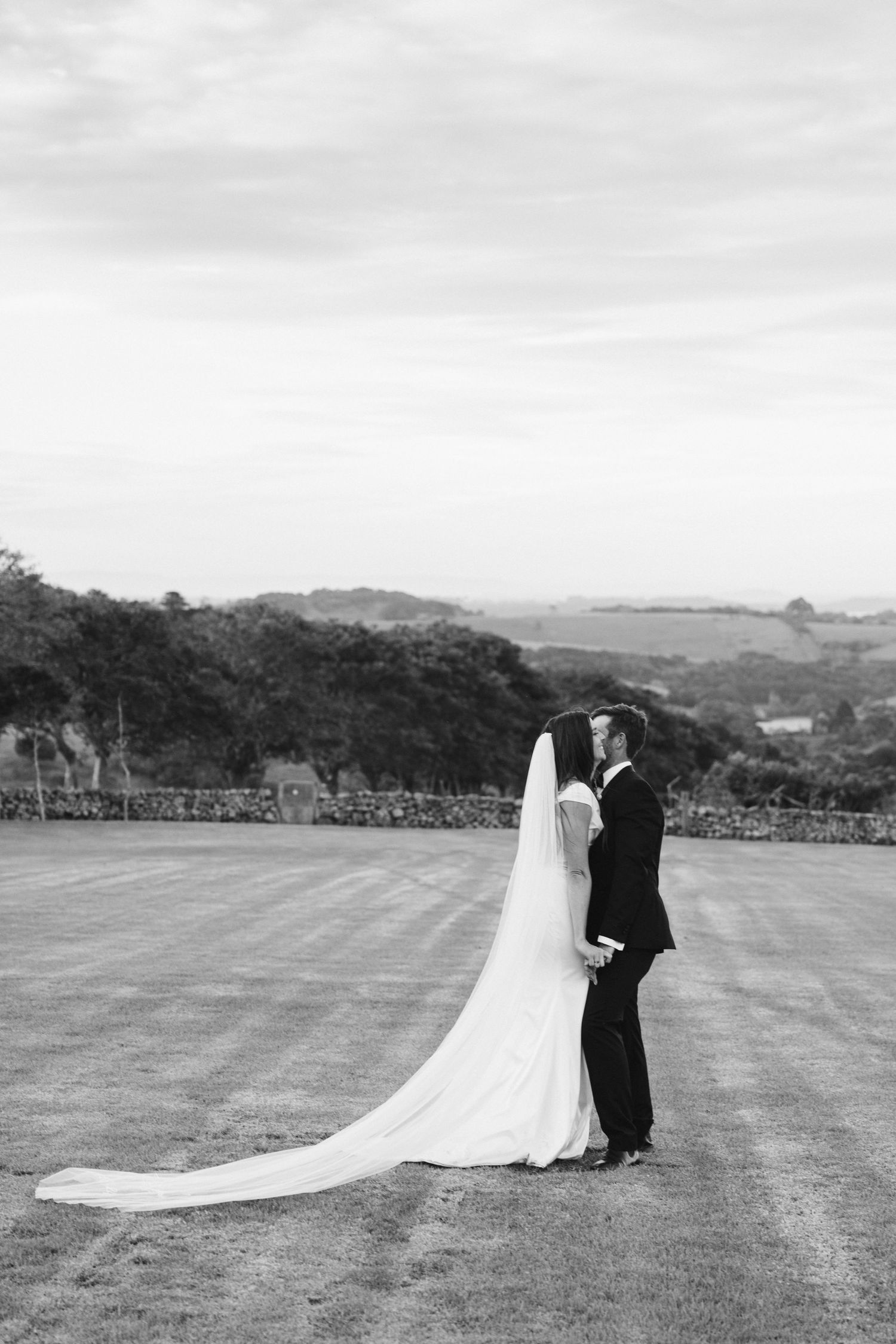 Black and white wedding portrait showcasing a long flowing veil against a countryside backdrop.