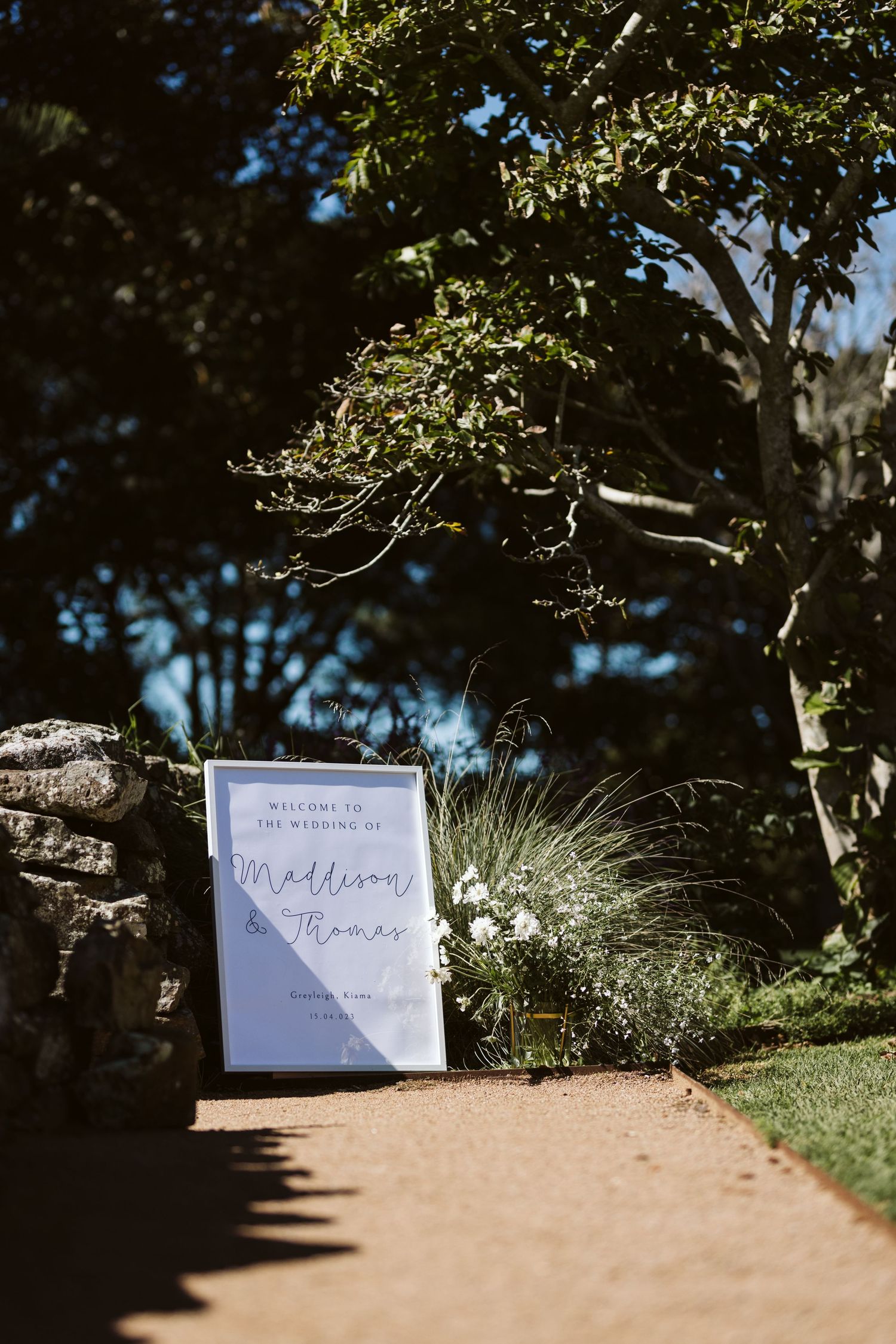 Wedding welcome sign displayed on path with tree and natural landscape in background.