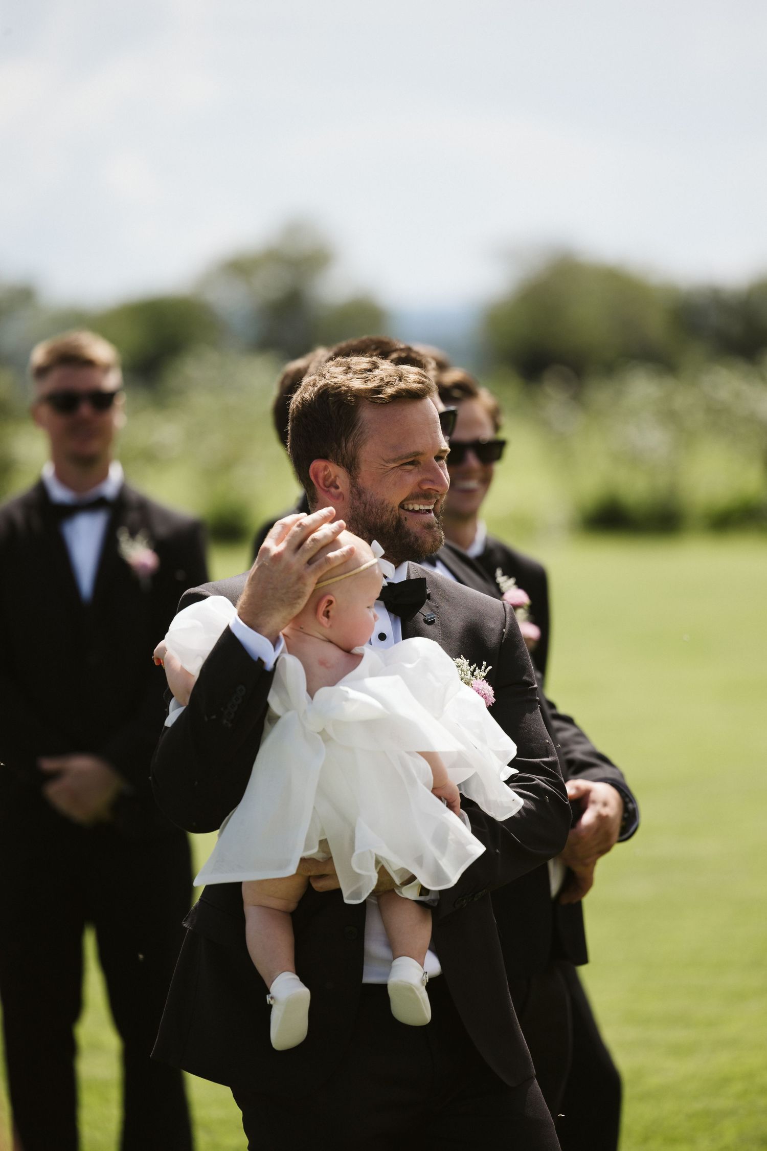 Wedding party member in tuxedo holds small child in white dress during outdoor ceremony.