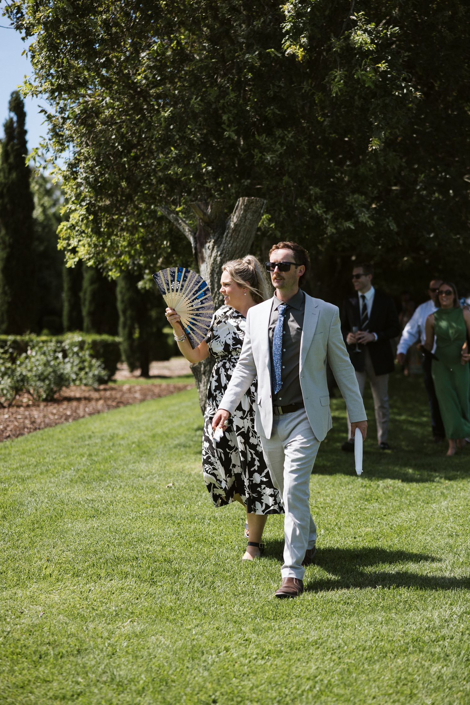 Wedding guests walking through a sunny garden setting with woman using decorative fan.