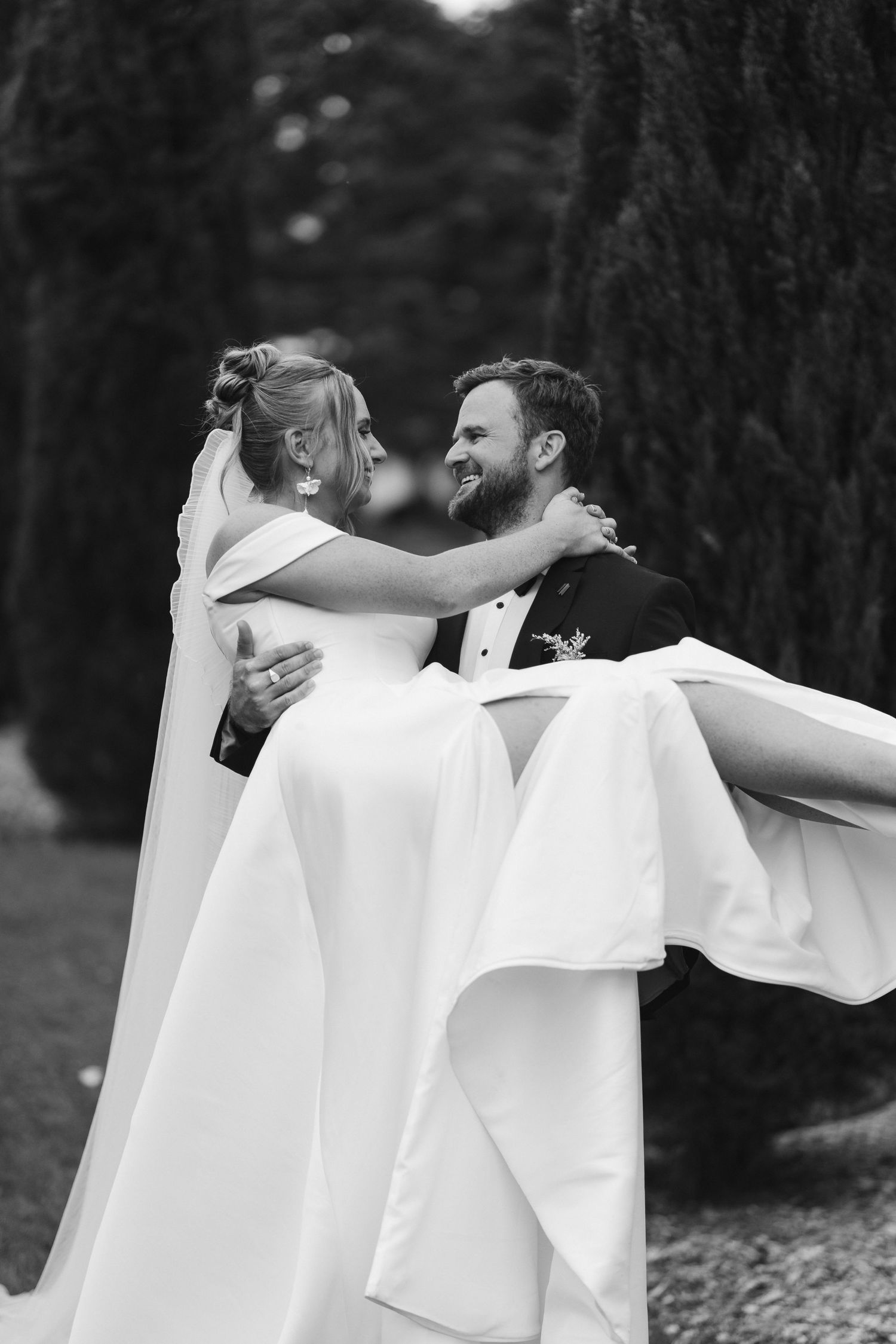 A romantic black and white wedding photo capturing a groom carrying his bride in a flowing white dress among trees.