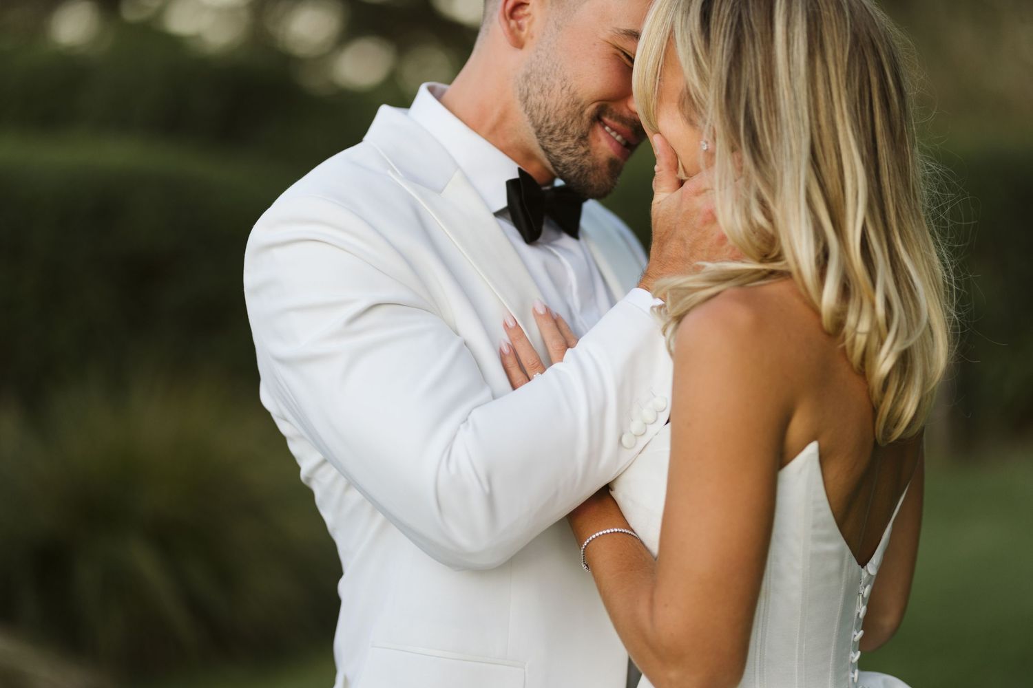 Intimate moment between bride and groom in white formal wear against garden backdrop.