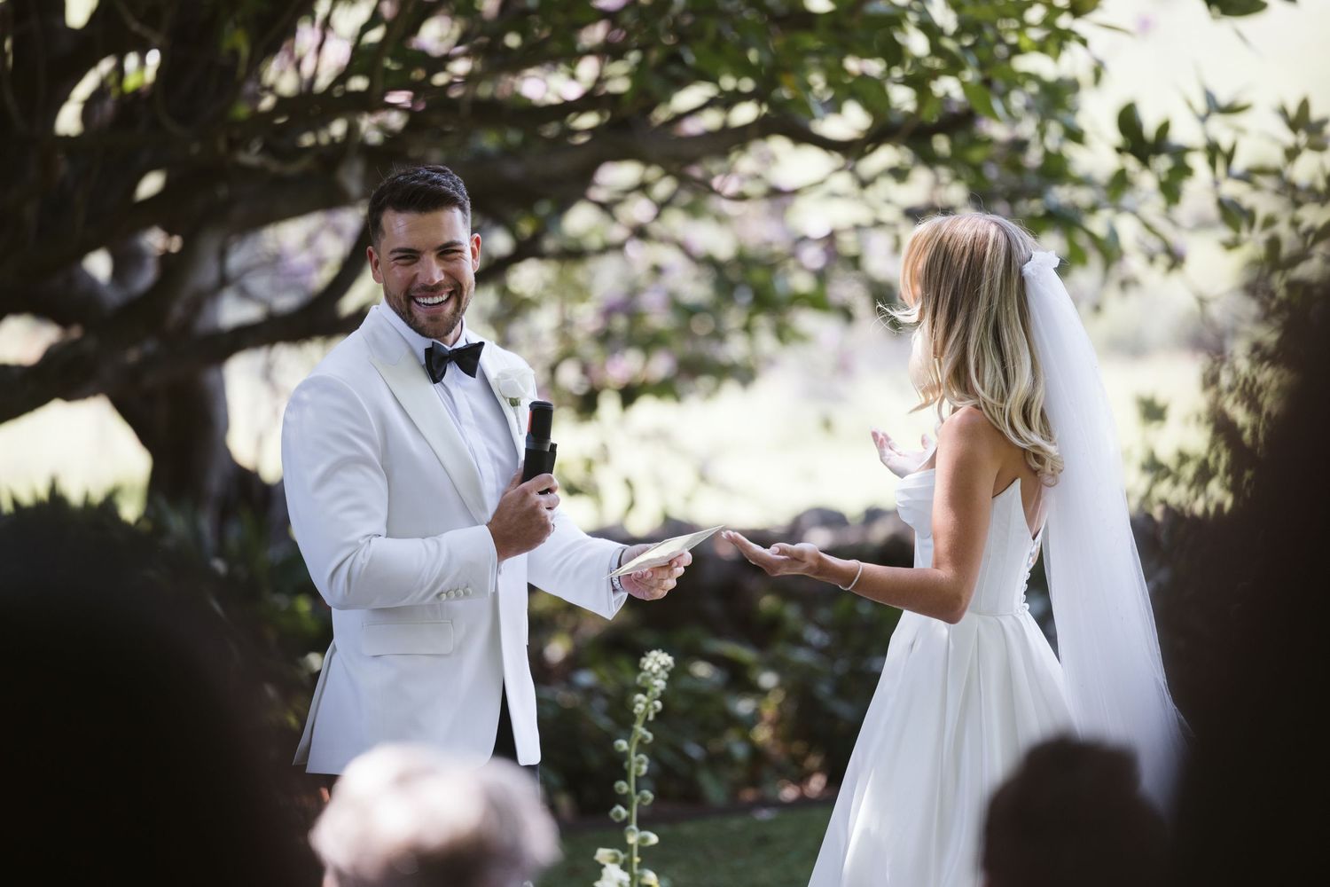 A couple exchanges wedding vows during an outdoor garden ceremony with sunlight filtering through trees.