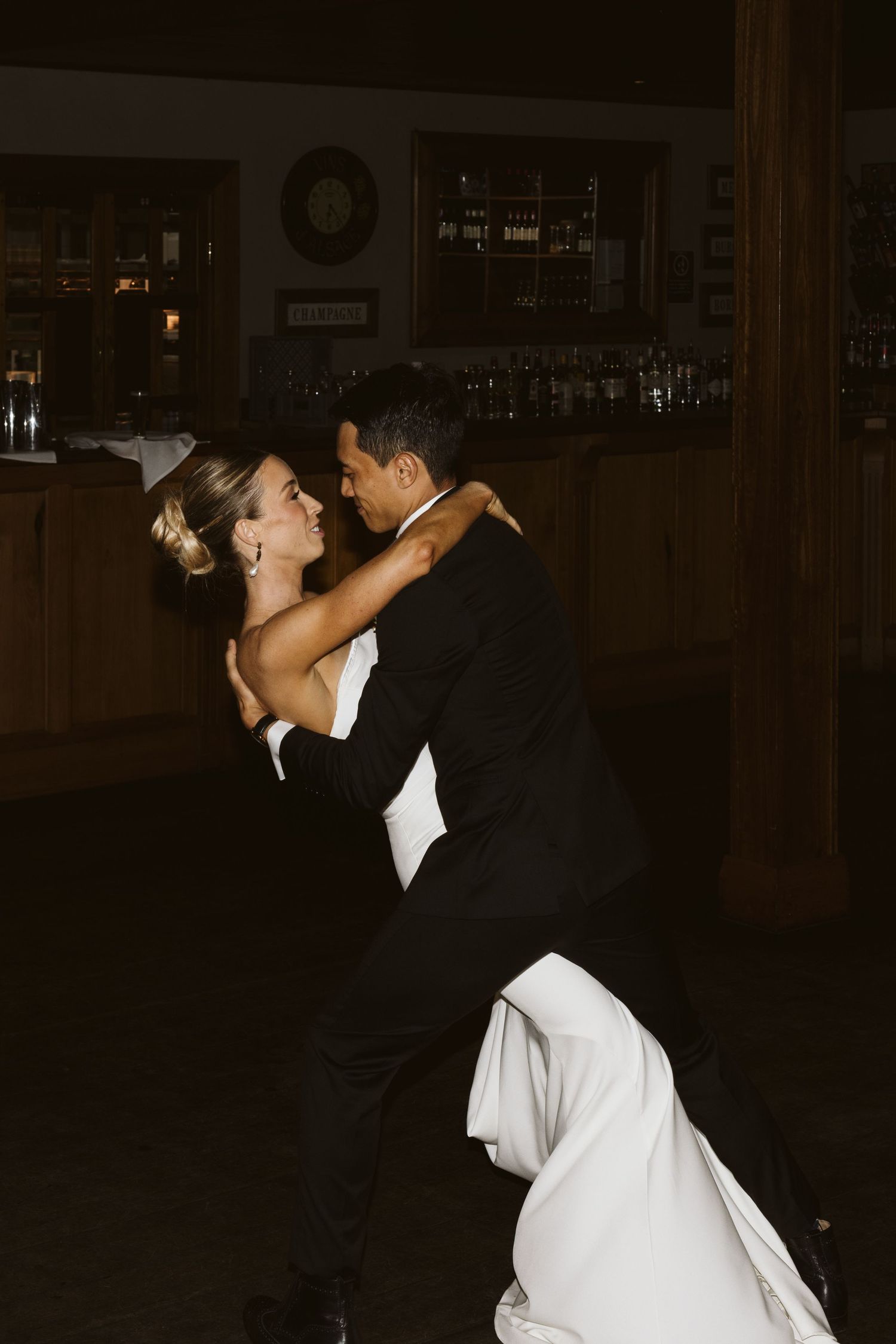 A couple shares a romantic dance dip during their wedding reception against a dark wooden bar backdrop.