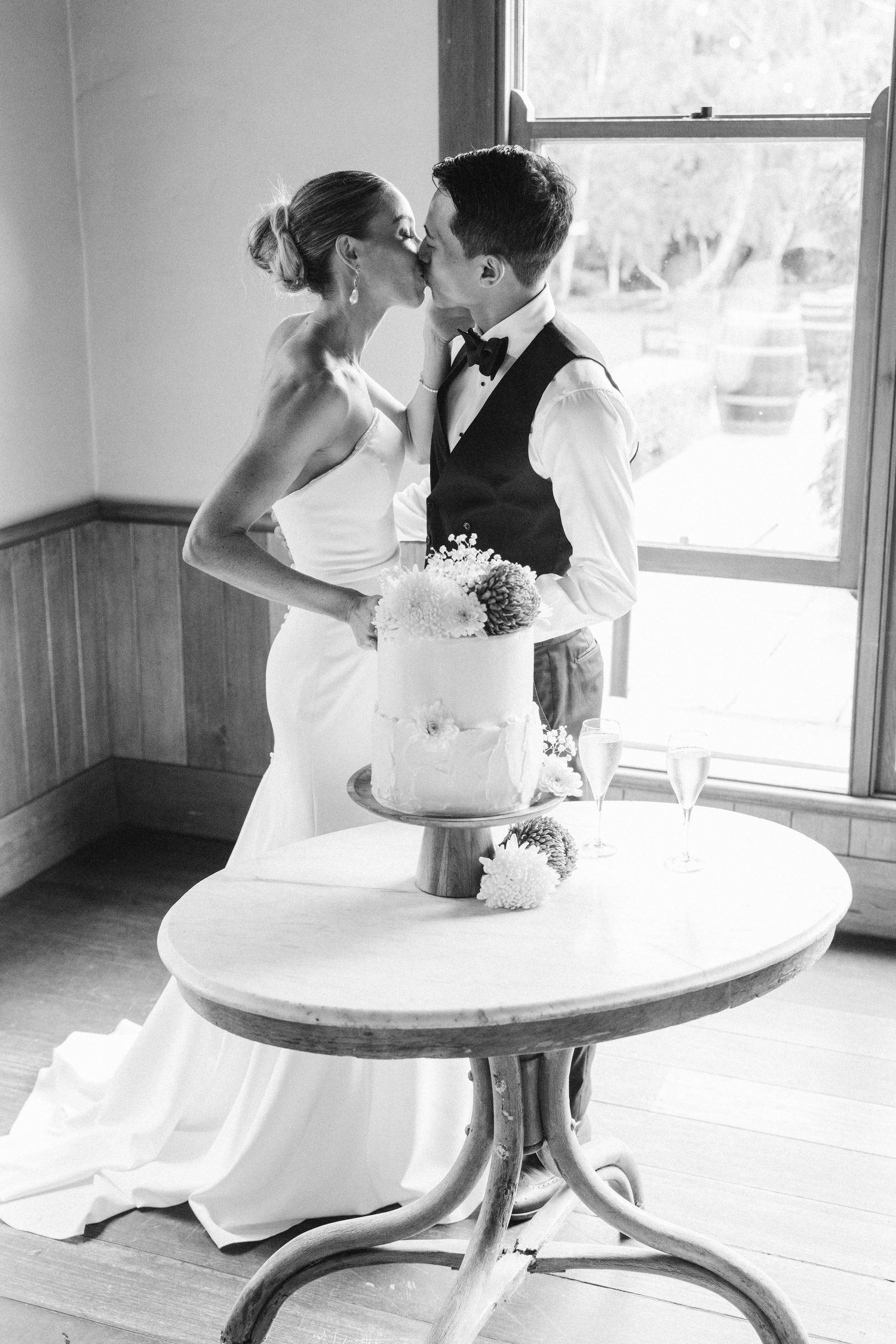 A couple shares an intimate moment beside their white tiered wedding cake near a sunny window.