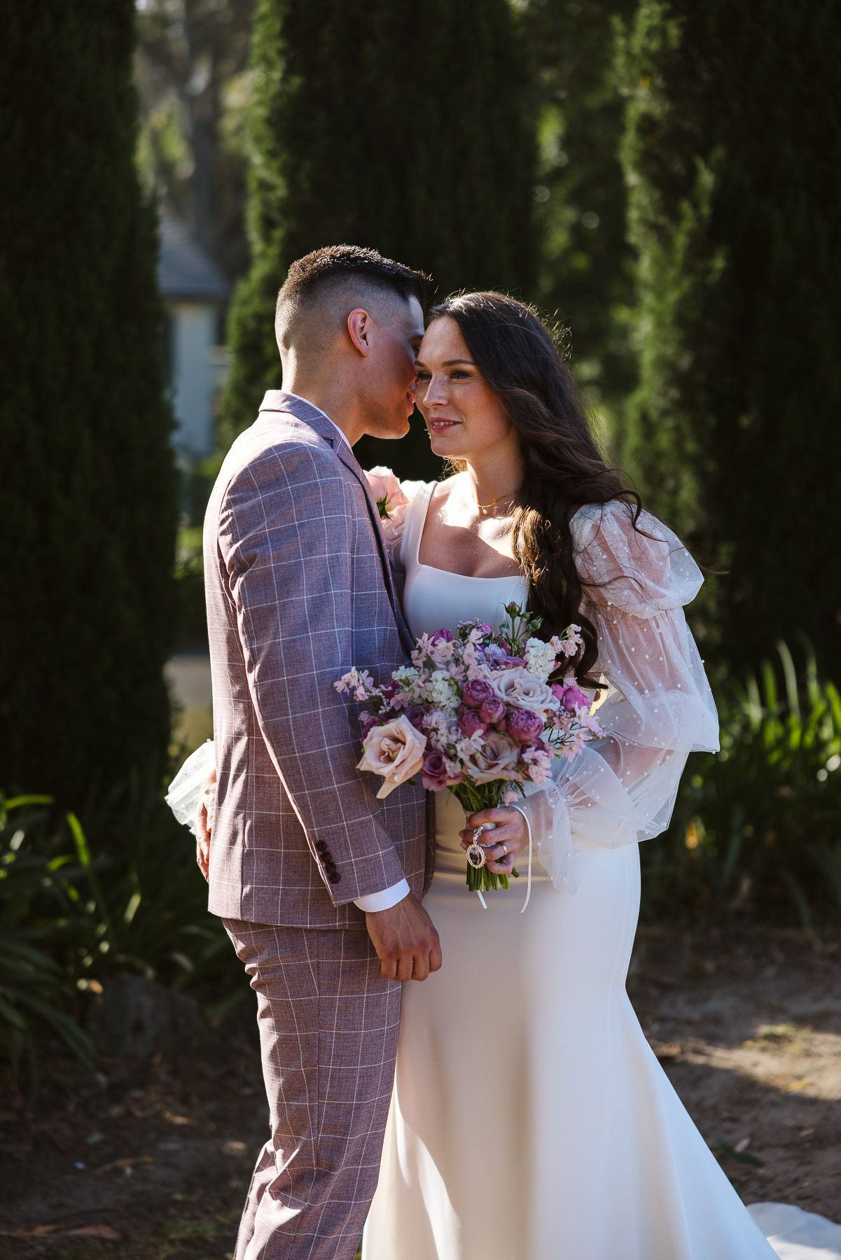 Bride and groom share a romantic moment outdoors, she holds a pastel bouquet in a white gown, he wears a plaid suit.