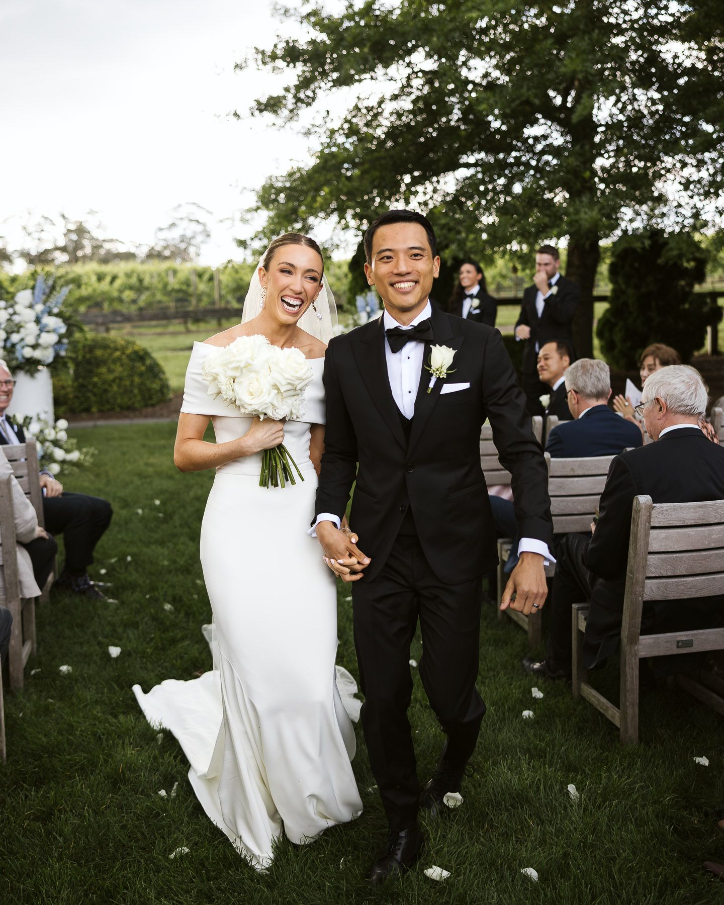 Happy newlyweds walk down outdoor wedding aisle, bride in white off-shoulder gown holding bouquet, groom in black tuxedo.