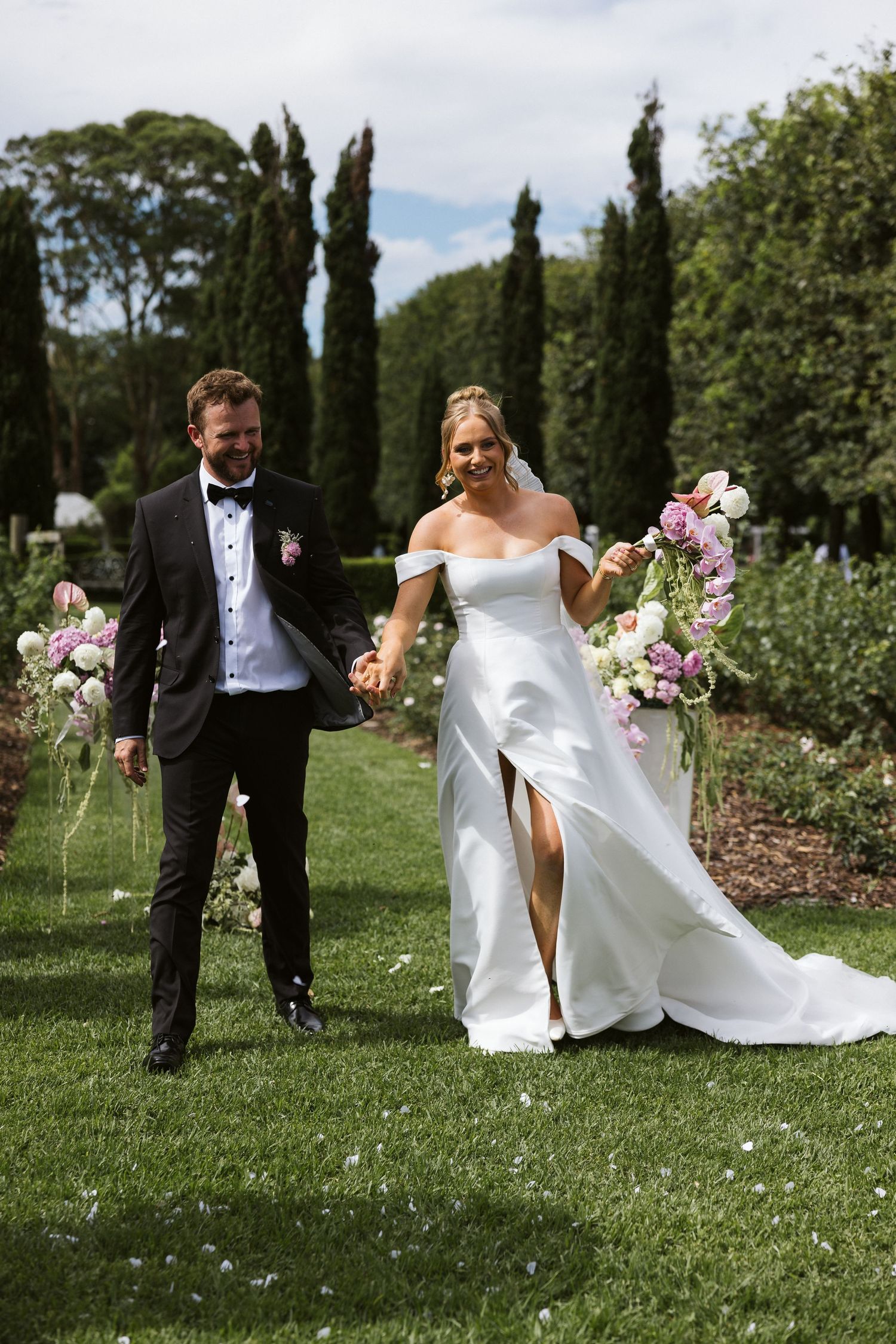 Newlyweds walk hand in hand through garden path with flowing white dress and tall cypress trees.