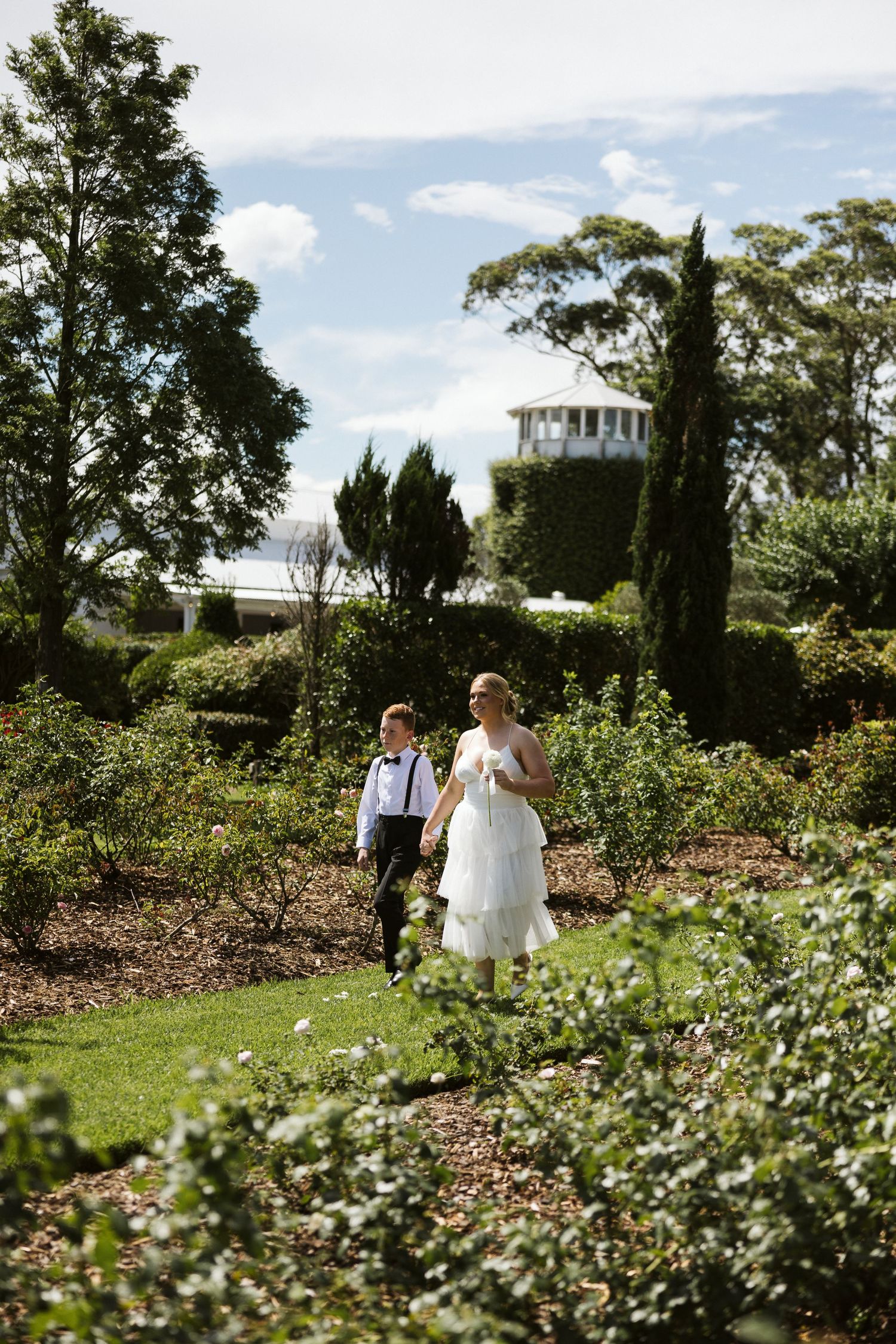 Wedding party members walk through a garden path with a white building visible in the distance.