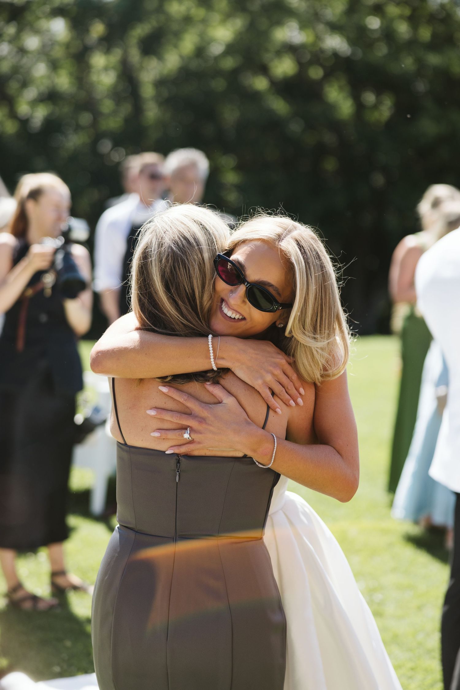 Two wedding guests share a joyful embrace on a sunny outdoor lawn during a celebration.