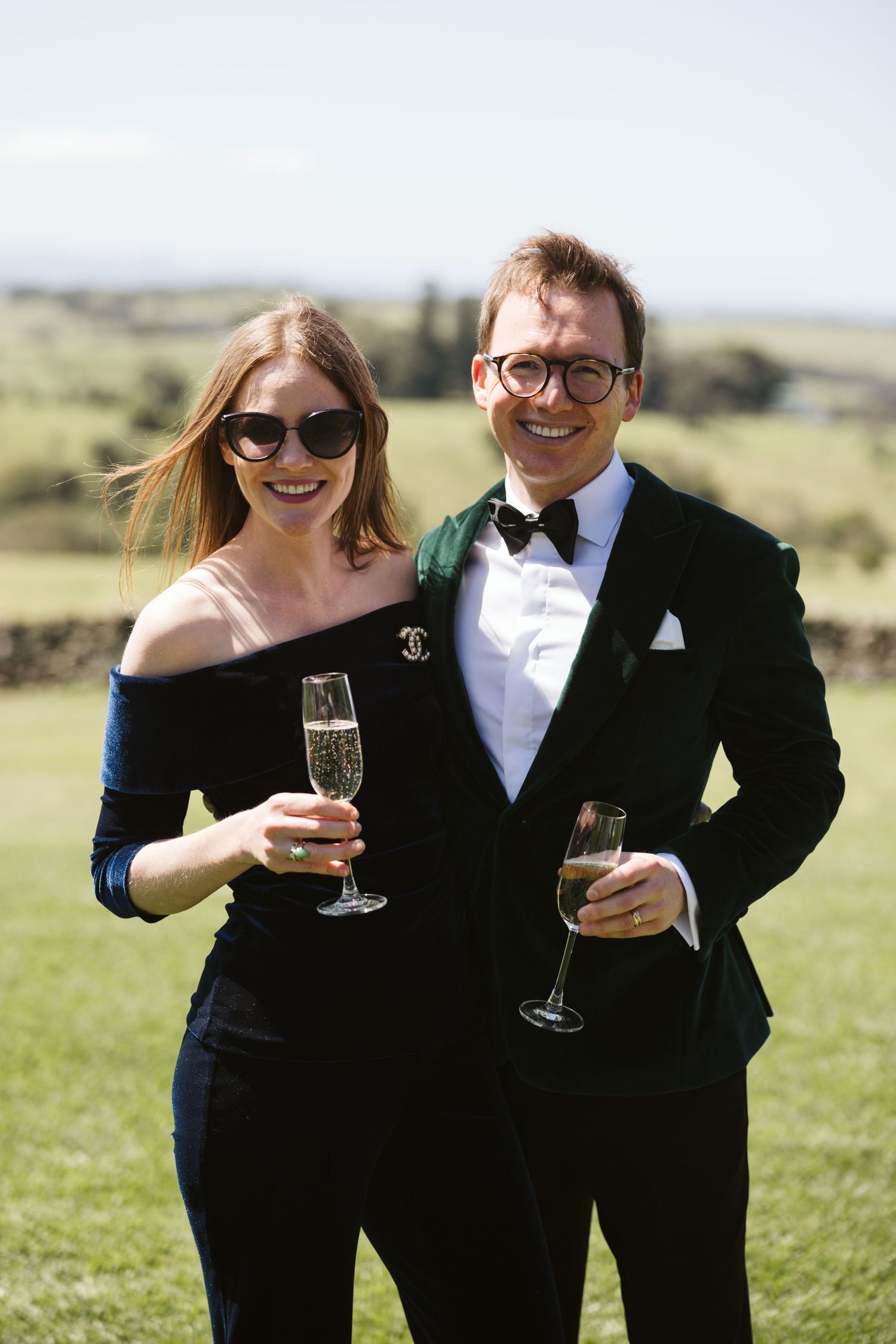 Elegantly dressed couple enjoying champagne outdoors in formal attire against a scenic landscape backdrop.