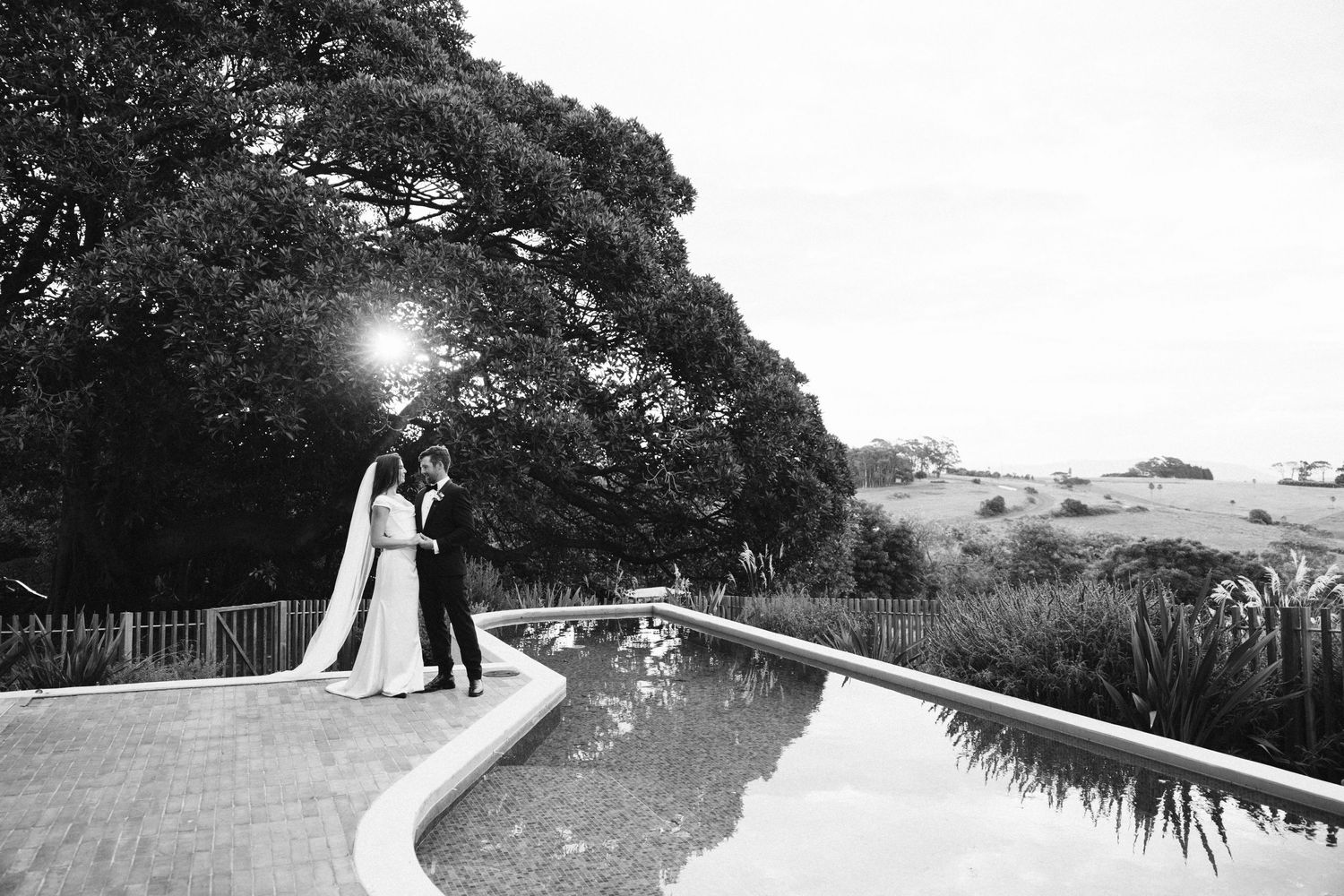 Silhouetted wedding couple standing by an infinity pool at sunset with rolling hills in background.