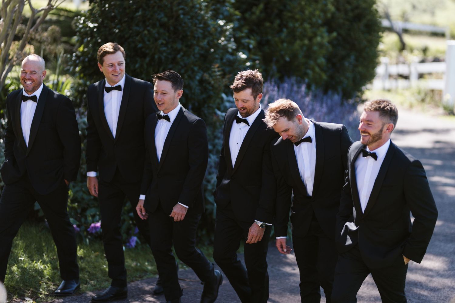 Wedding party in tuxedos walking together along outdoor garden path.