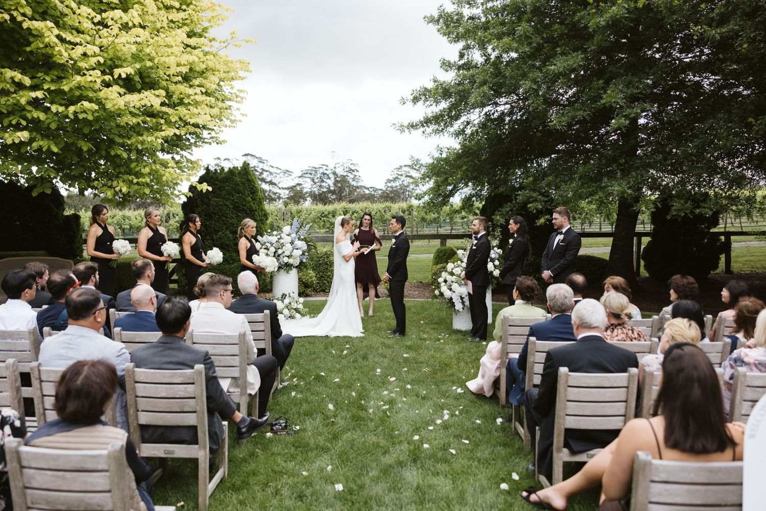 Outdoor wedding ceremony on a lush green lawn with white chairs and floral arrangements under trees.