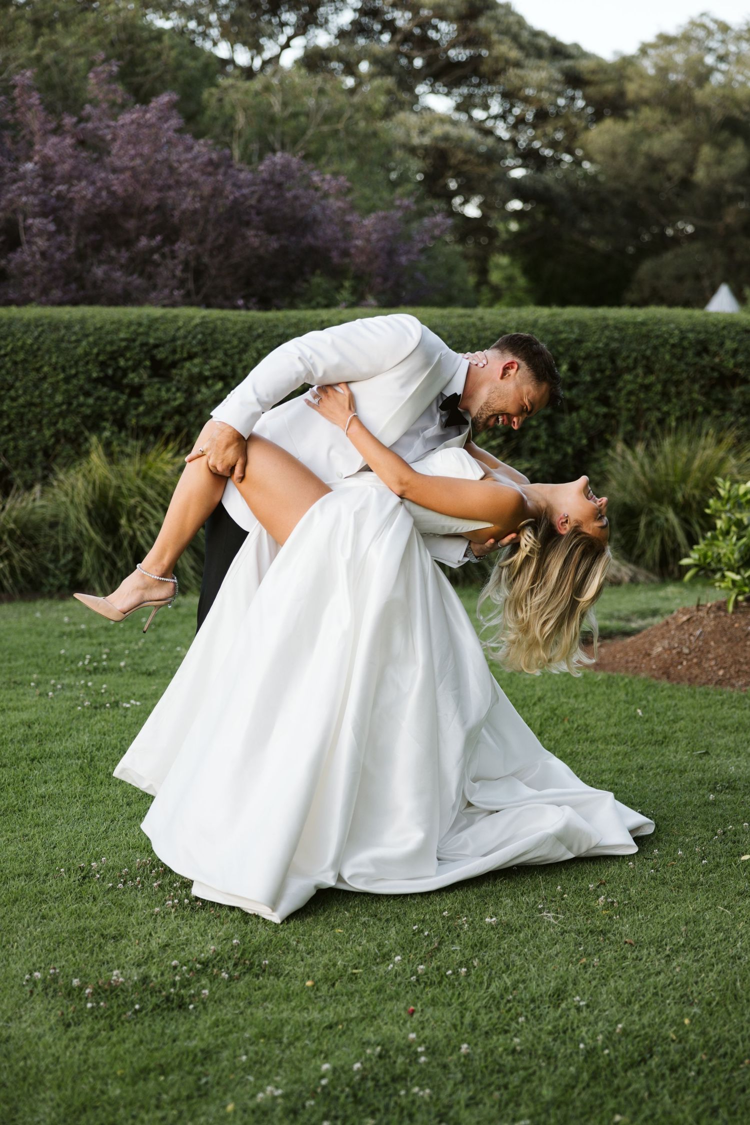Playful dip pose of newlyweds on garden lawn in formal attire.