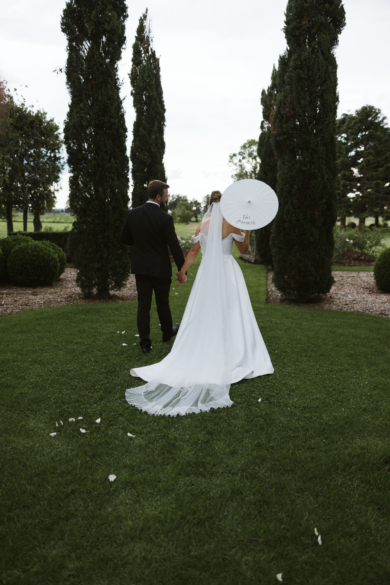 Couple holding white parasol walks through garden with tall cypress trees.