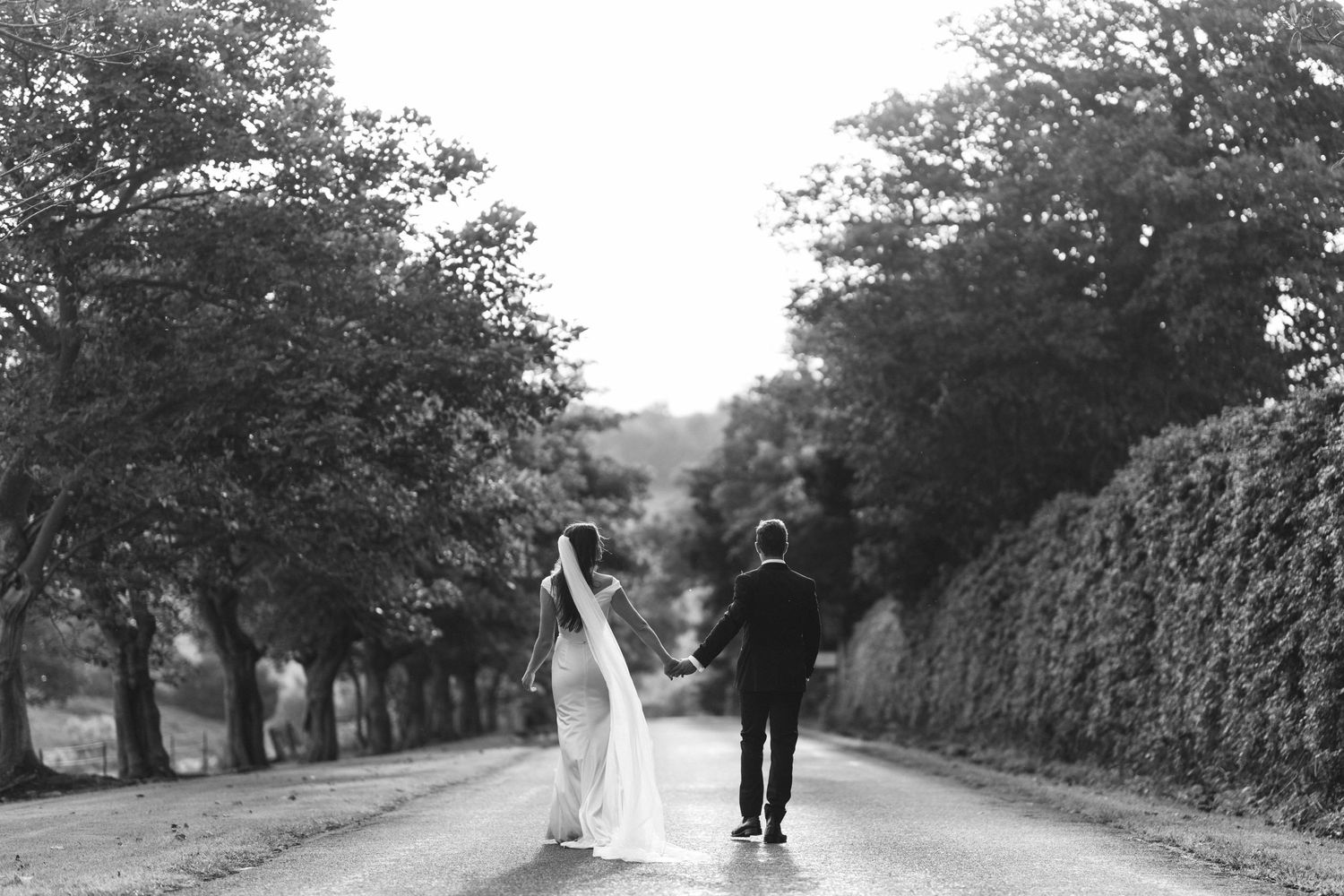 A wedding couple walking hand in hand down a tree-lined country road in black and white.