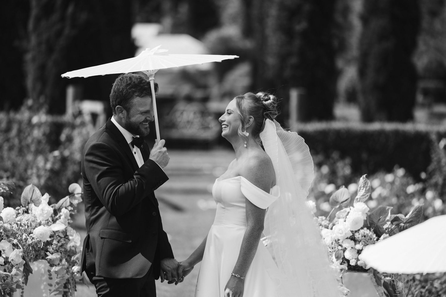 A playful black and white wedding photo with a parasol in a garden setting.
